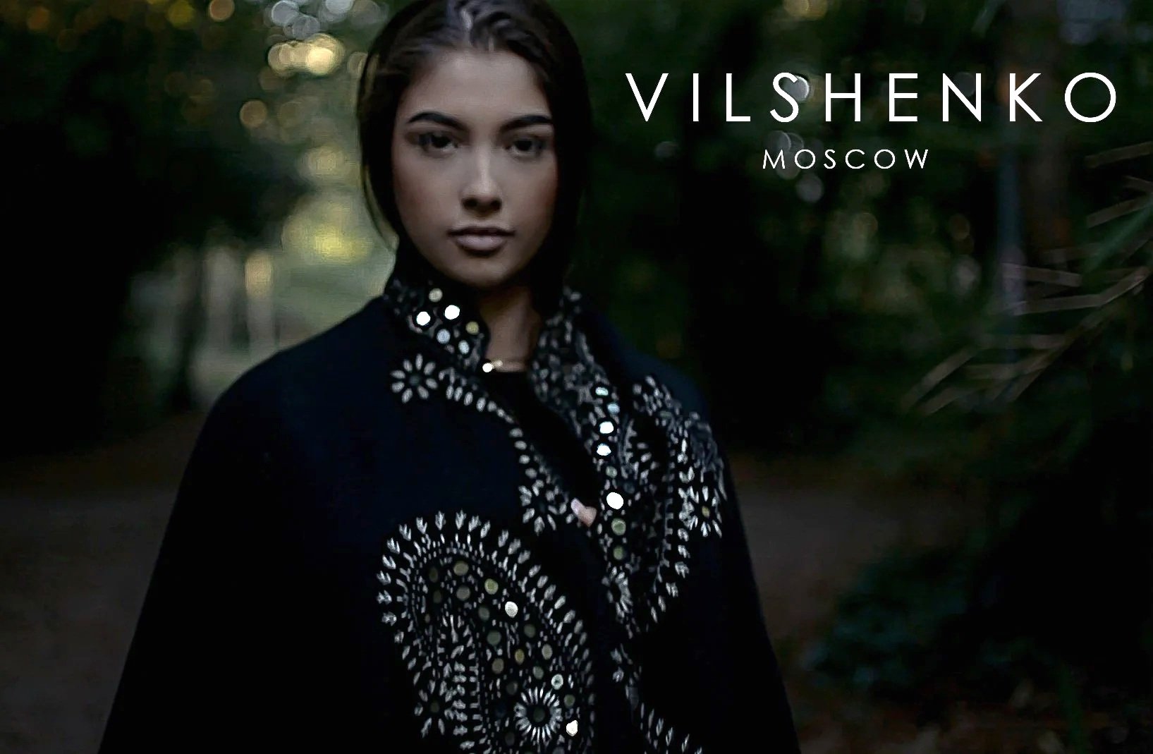 A woman with dark hair wearing a black embroidered jacket in a forested outdoor setting at dusk.