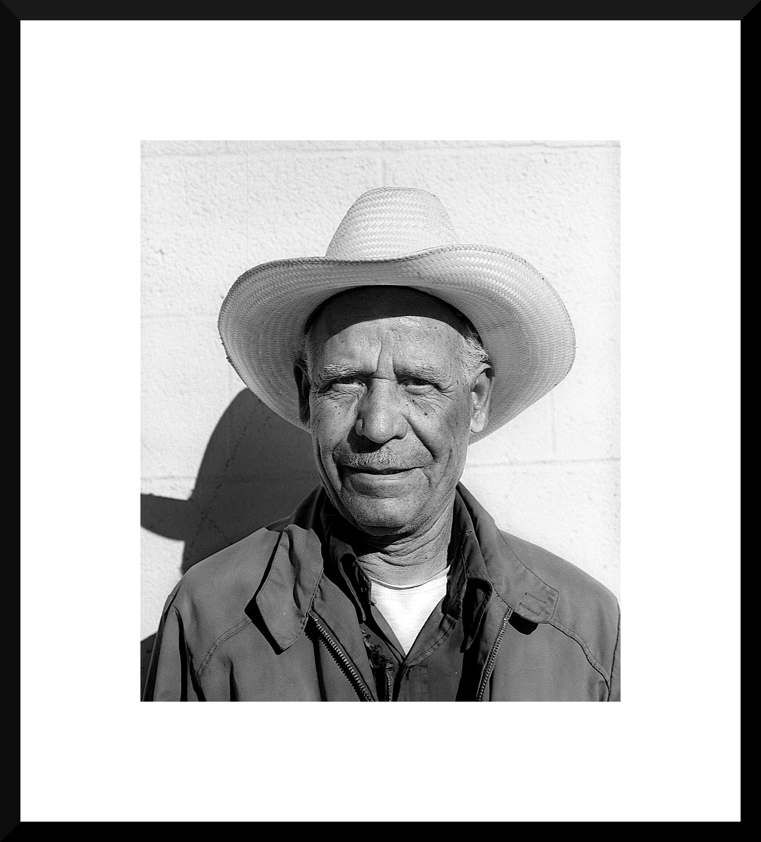 Black and white photo of an older man wearing a wide-brimmed straw hat, smiling slightly, with a shadow cast on a light-colored wall behind him.