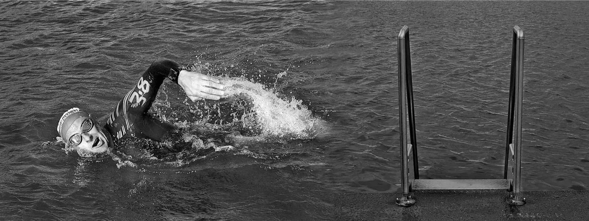 Swimmer wearing goggles and a swim cap, swimming in open water with a ladder on the side.