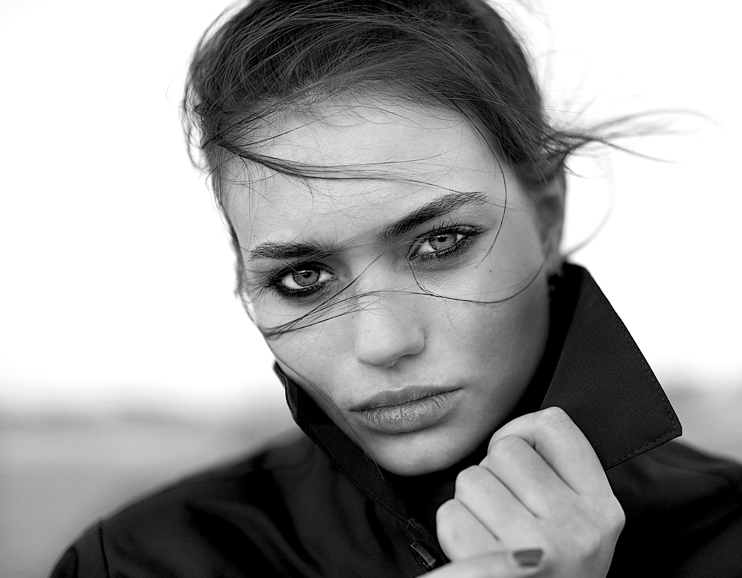 Black and white close-up portrait of a woman with wind-blown hair, intense gaze, and holding the collar of her jacket with her hand.