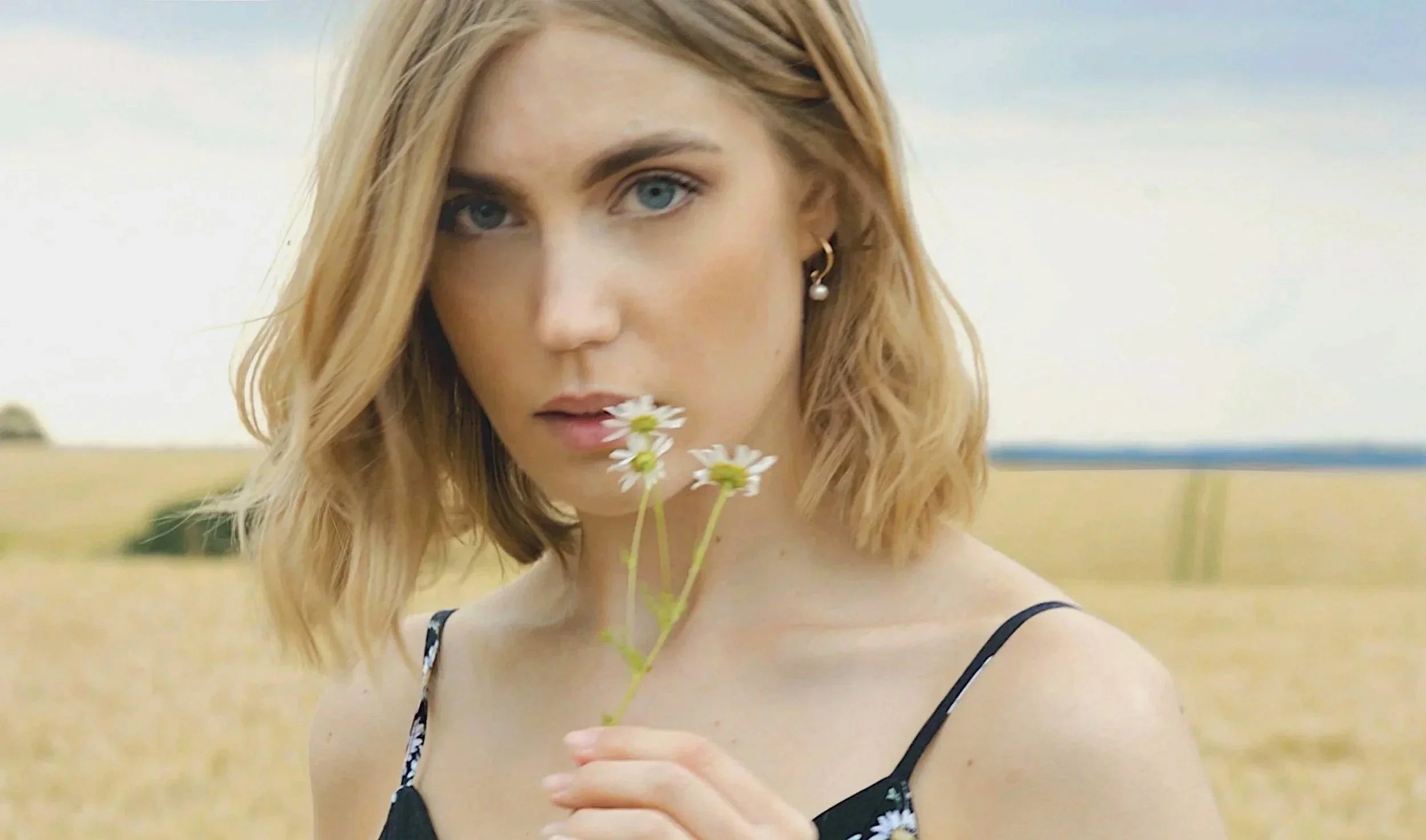 A young woman with blonde hair and blue eyes holding a small bouquet of daisies in front of her face outdoors.