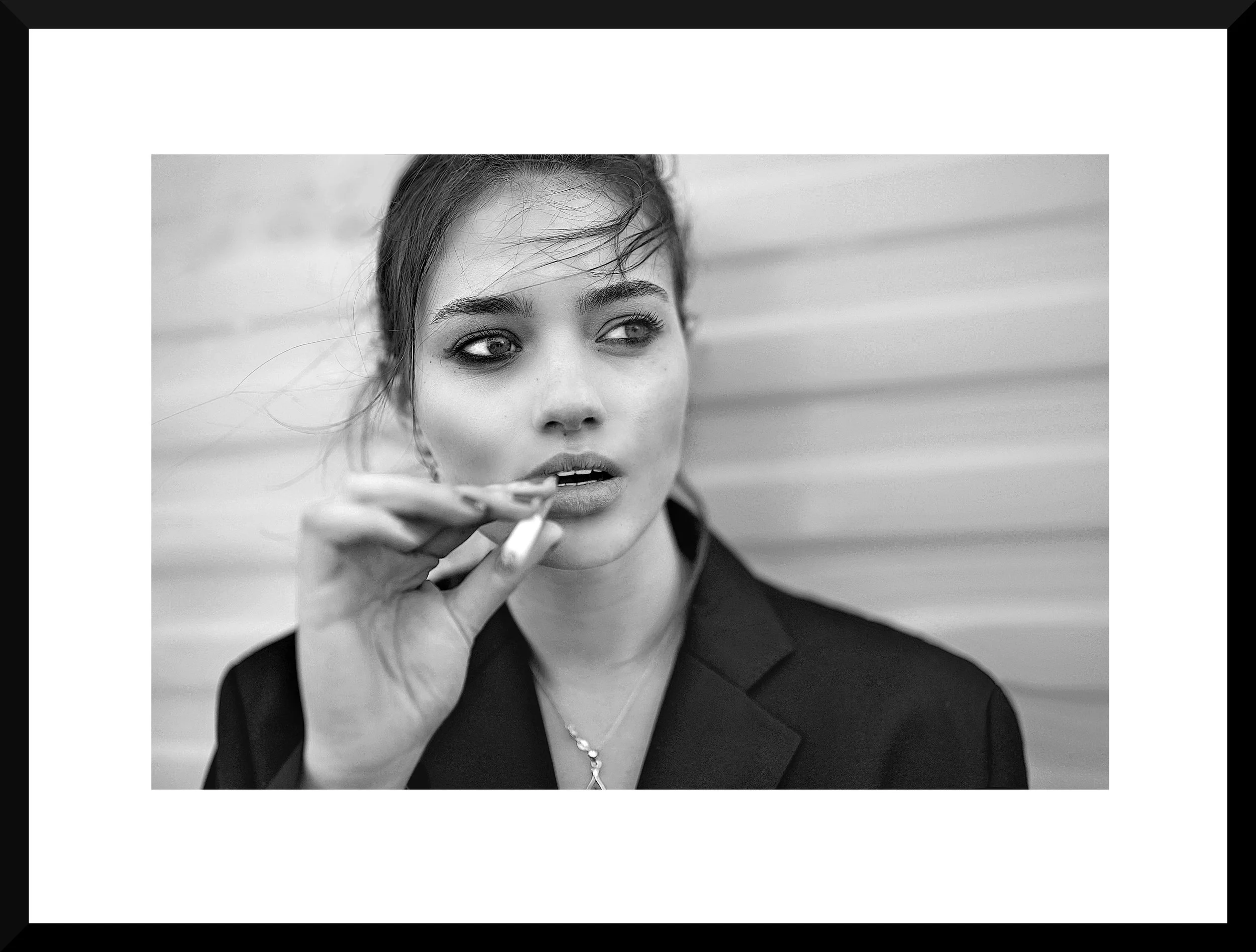Black and white photo of a young woman with her hair pulled back, wearing a dark blazer and earrings, holding a cigarette near her lips, standing against a horizontal wall.