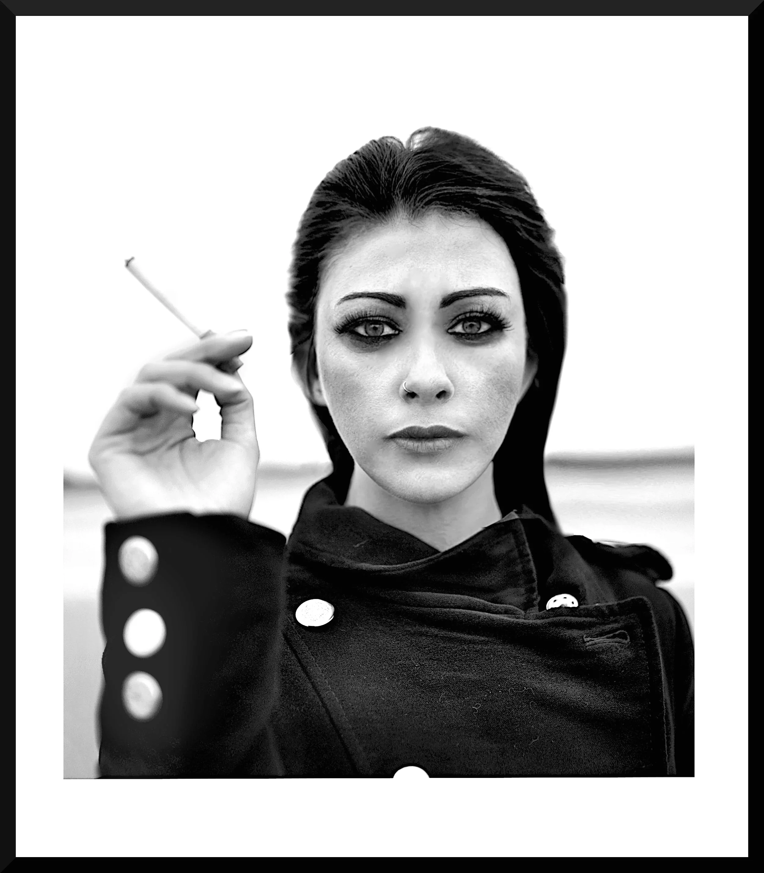 Close-up of a woman with dark hair and piercing eyes, holding a cigarette near her face, wearing a black outfit with white polka dots on the sleeve, black lipstick, and a nose ring.