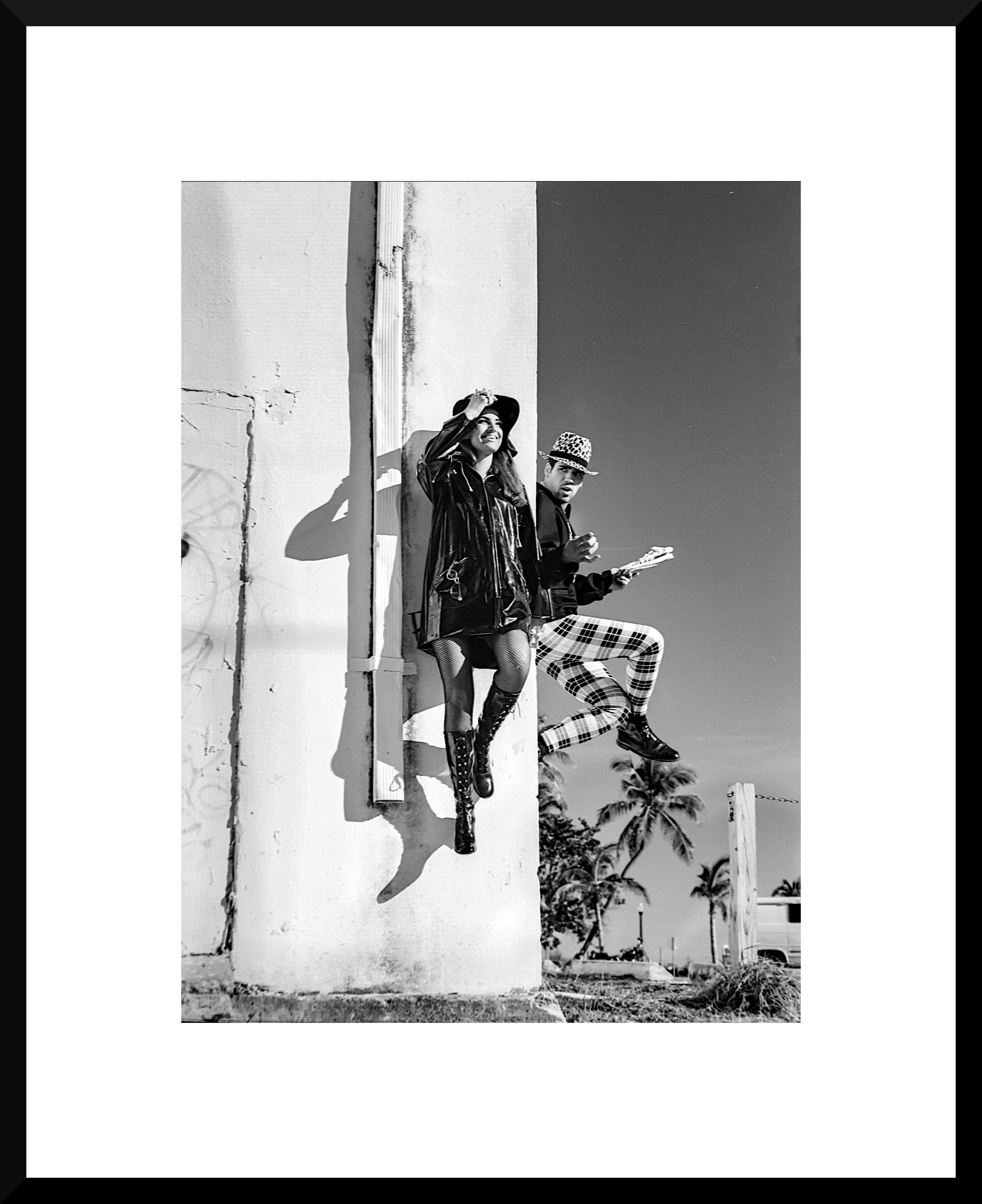 Black and white photo of a woman and a man sitting on a wall or ledge outdoors, with palm trees in the background. The woman is smiling, wearing a hat, jacket, and boots, while the man is holding a newspaper or magazine, wearing plaid pants and a hat.