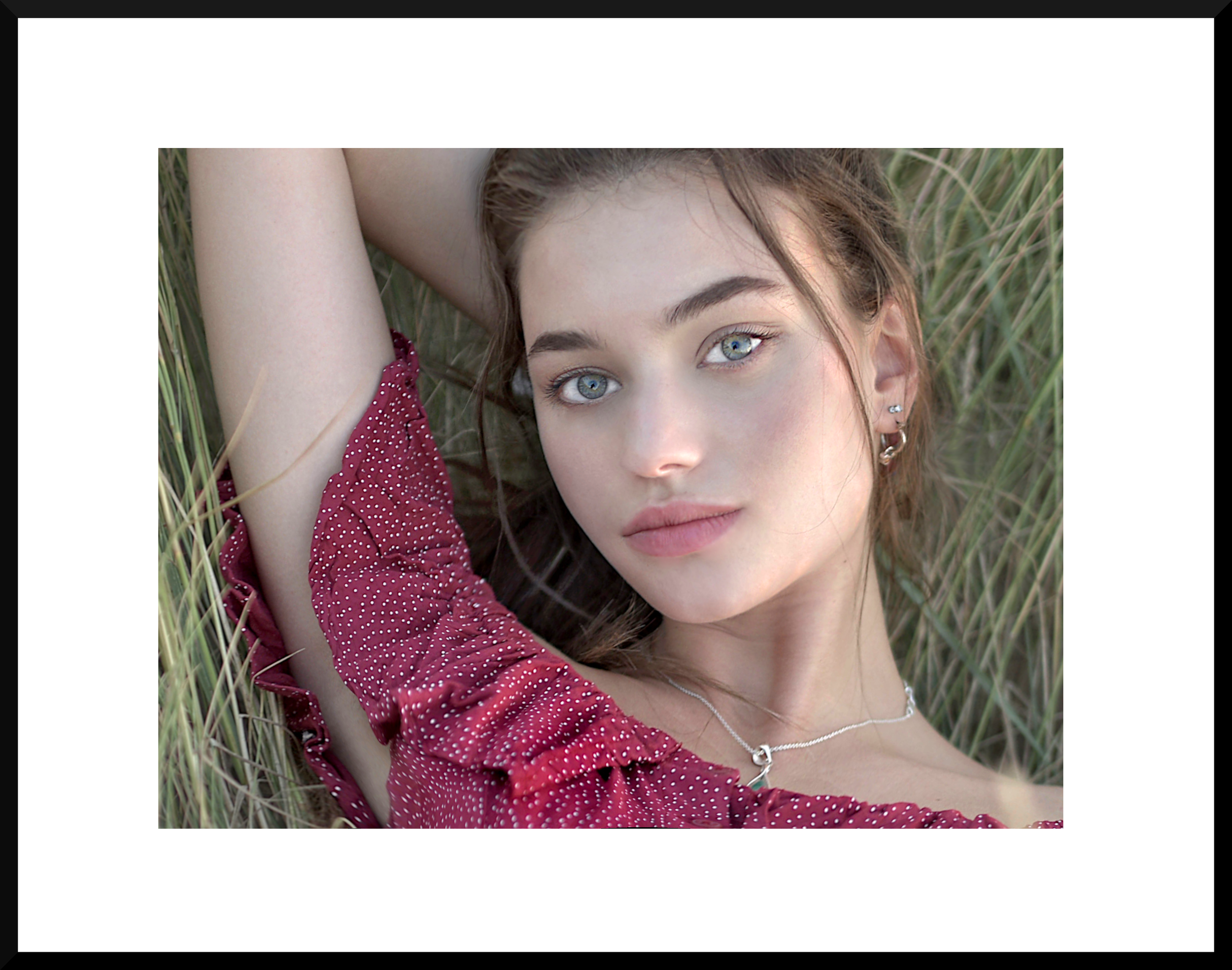 Close-up of a young woman with blue eyes lying in tall grass, wearing a red dress with white polka dots and silver jewelry.