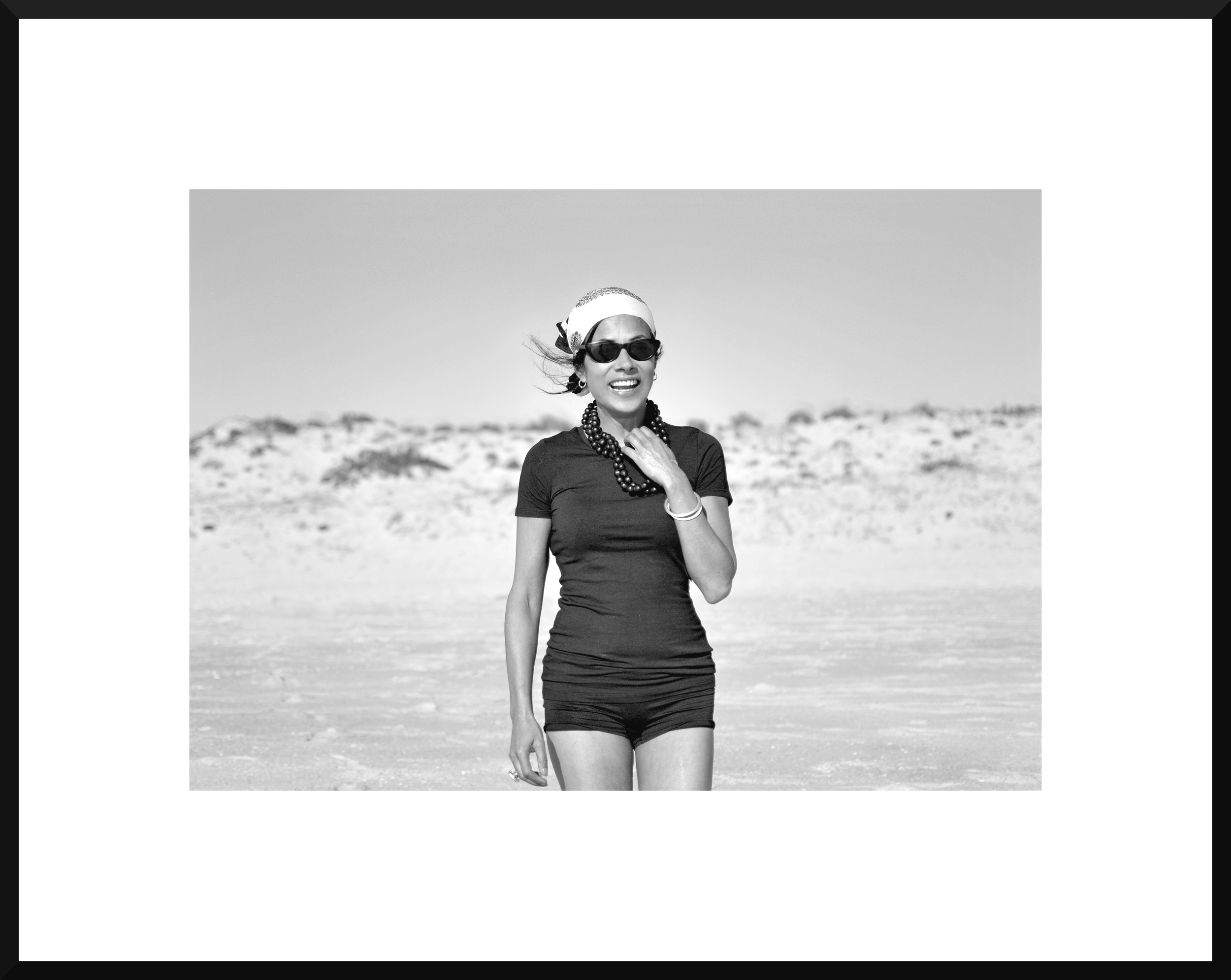 Black and white photo of a woman smiling on the beach, wearing sunglasses, a head scarf, a necklace, and a t-shirt with shorts.
