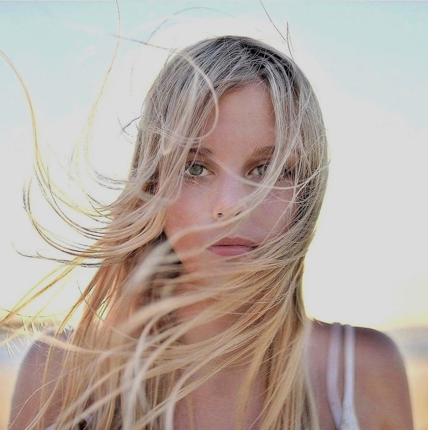 Close-up of a young woman with long, wavy blonde hair and blue eyes, outdoors with windy weather.