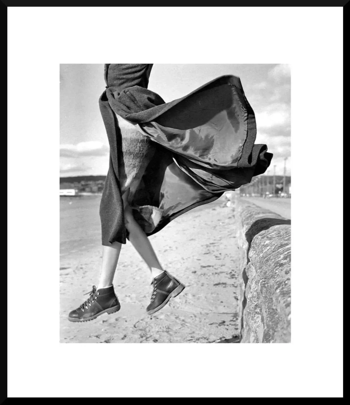 Black and white photo of a person jumping over a low stone barrier near a body of water, wearing sneakers, with a scarf or cape flowing in the wind, against a cloudy sky.