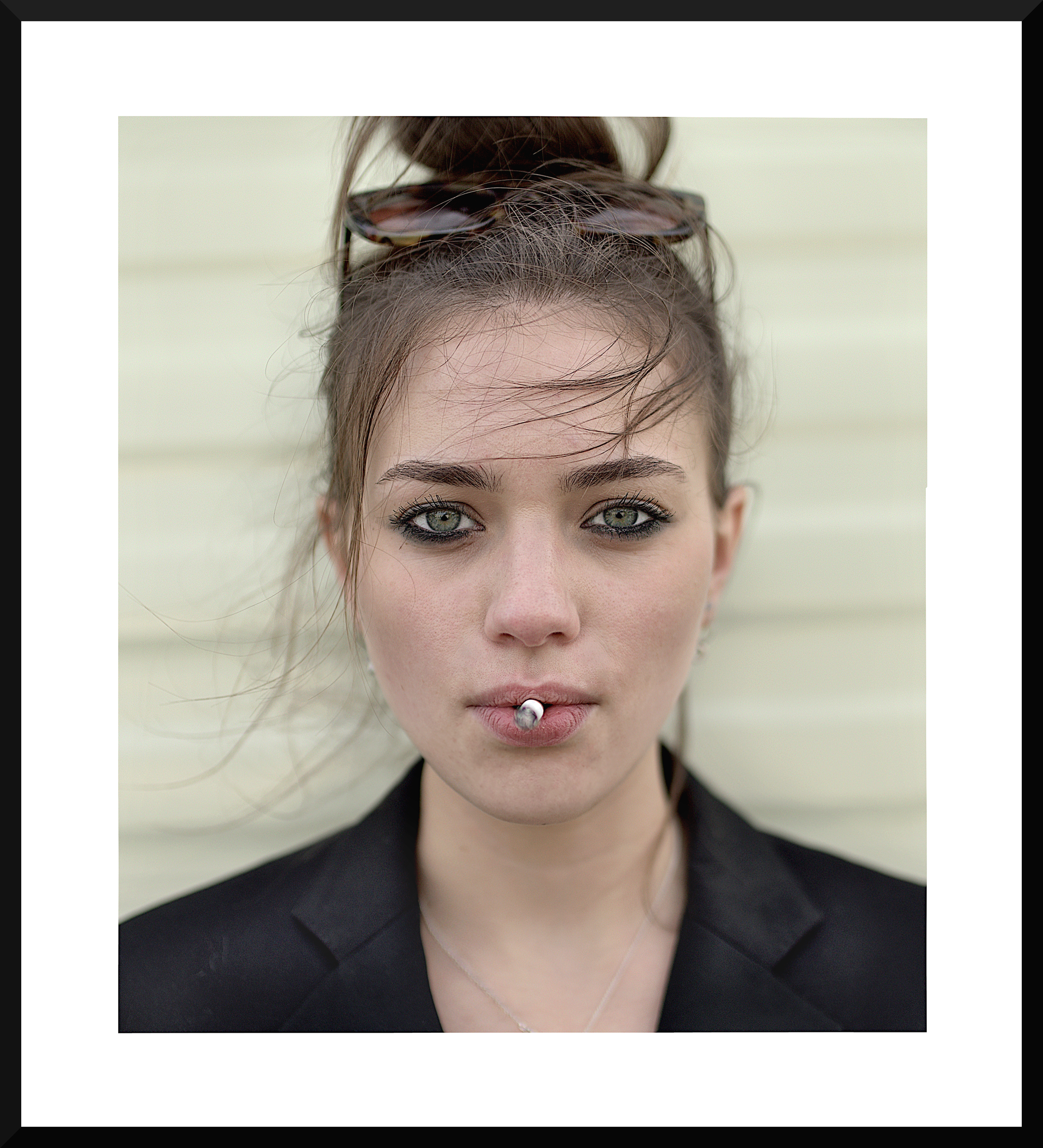A young woman with green eyes and dark makeup, smoking a cigarette, with sunglasses resting on her head and her hair tied in a messy bun, in front of a light-colored background.