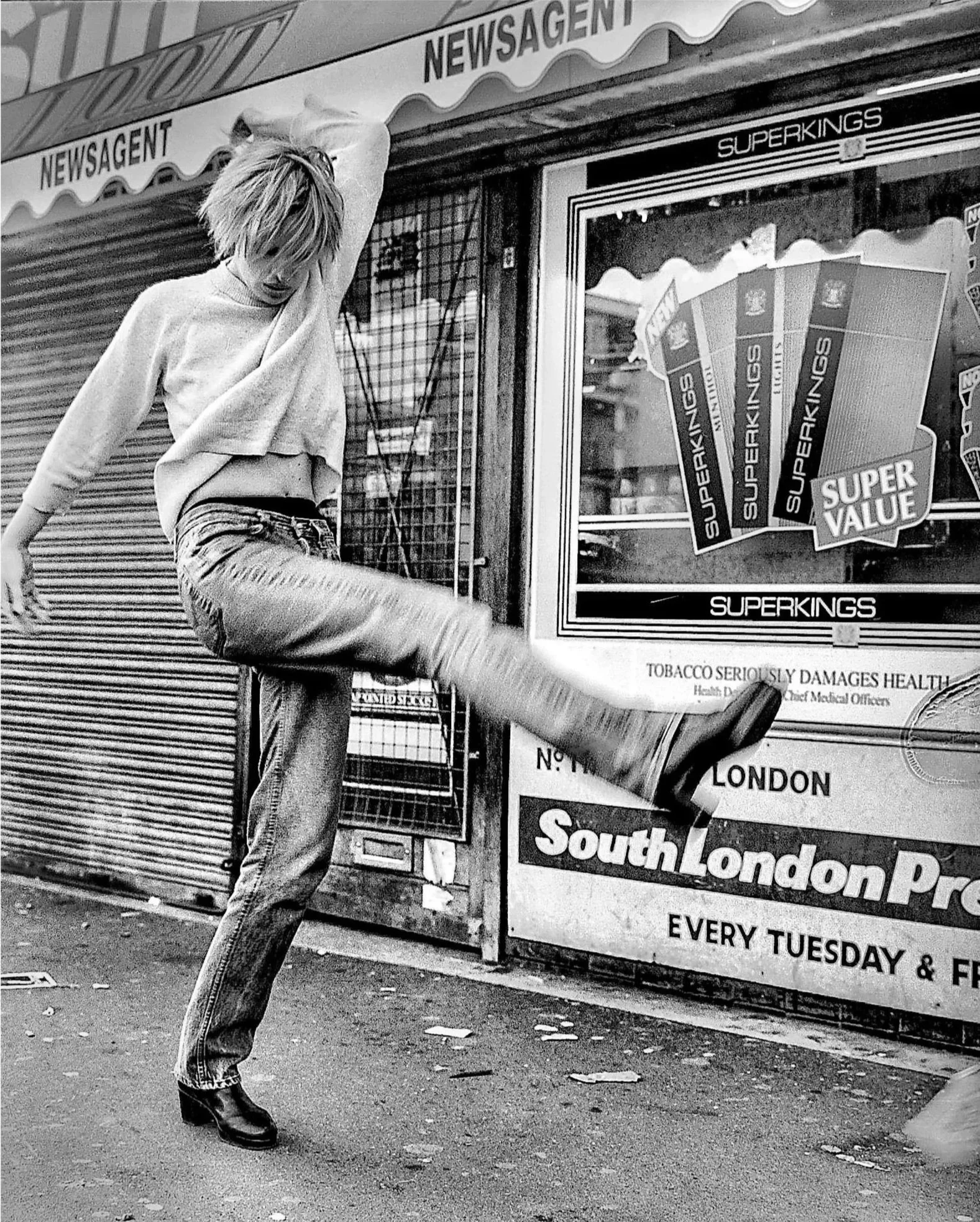 A young man with messy blonde hair, wearing a loose shirt, jeans, and boots, standing in front of a closed shop with signs promoting Superking cigarettes and a health warning about tobacco damaging health. He is stretching one arm overhead and has one foot lifted, leaning against a shuttered storefront.