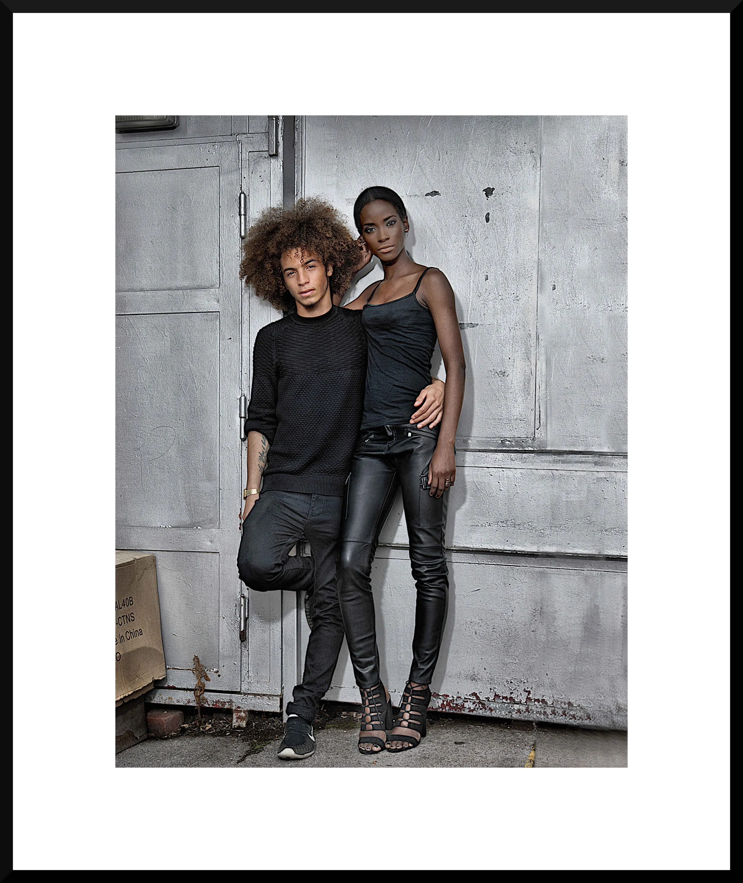 Two young adults, a man and a woman, standing against a gray, industrial wall. The man has curly hair, wearing a black sweater and pants, with one knee up against the wall. The woman has straight hair, wearing a black tank top and leather pants, with her arm around the man's shoulder.