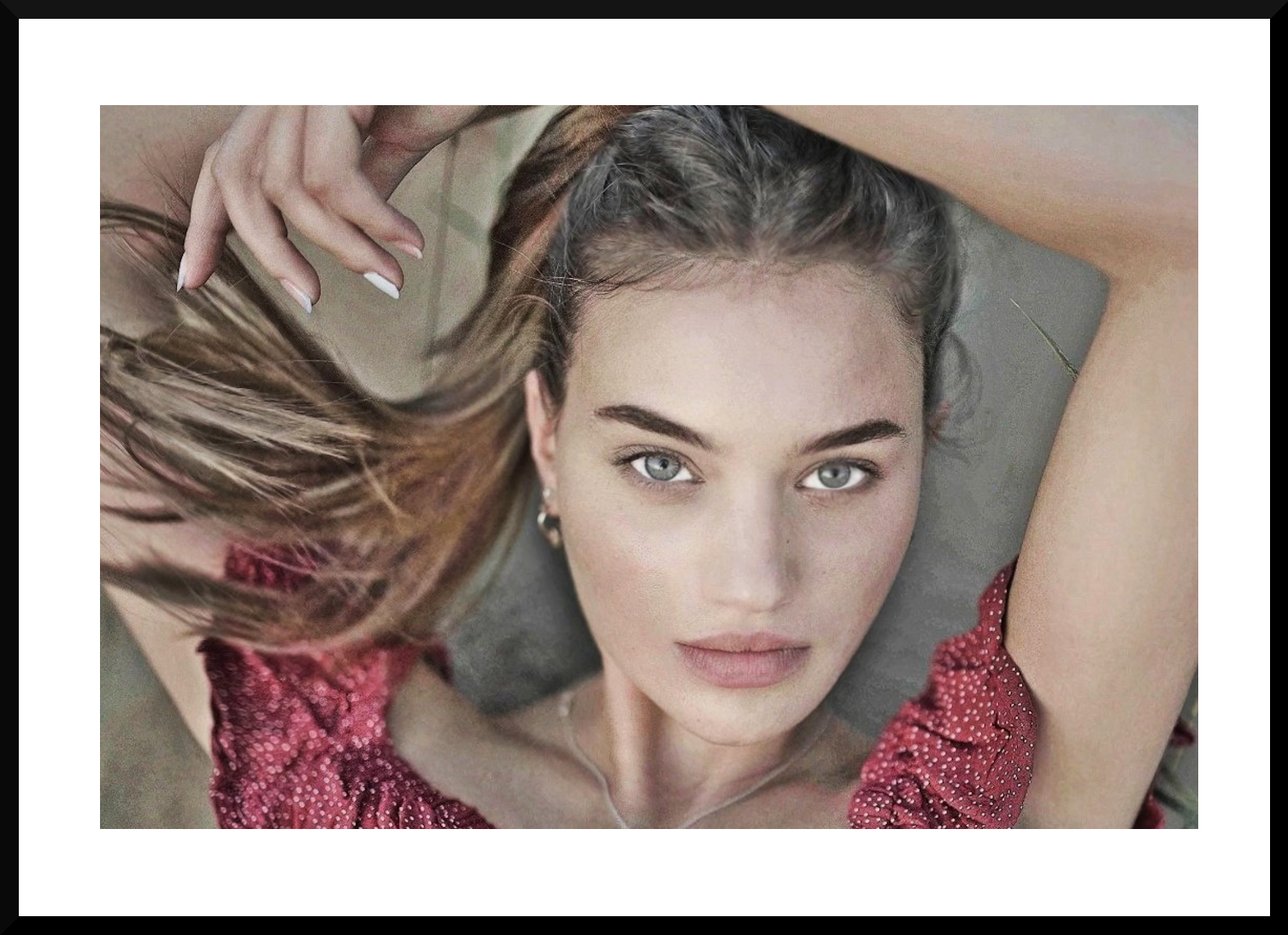 A young woman with striking blue eyes and light makeup lying on her back, looking directly at the camera, with one arm resting above her head and her long brown hair spread out to the side.