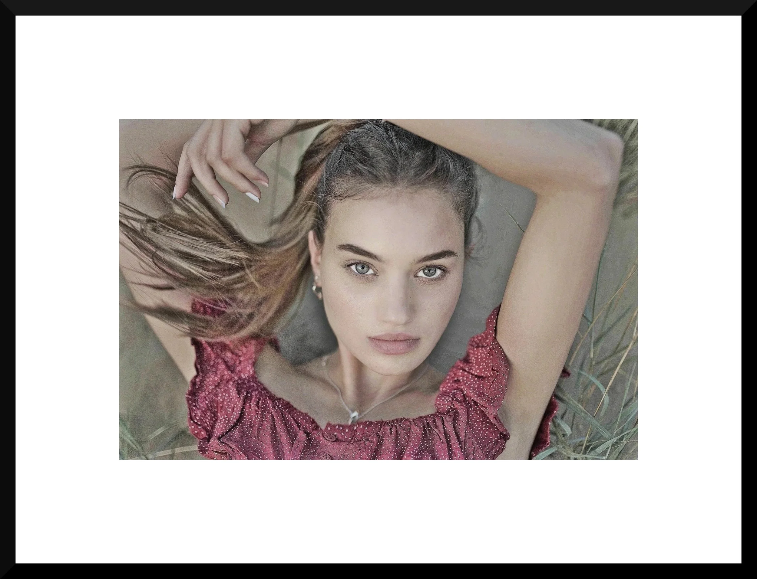 A young woman with light skin and blue eyes lying on her back in a field of grass, wearing a red dotted dress, with her hair spread out and one hand touching her hair.