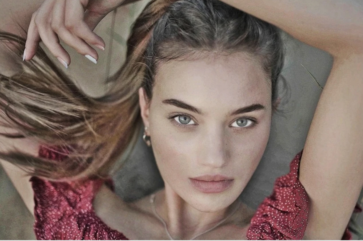 A woman with blue eyes and light skin lies on her back, her head tilted back, with one hand holding her hair. She is wearing a red patterned top and has earrings and a necklace. The photo is taken from above, showing her face and part of her arms.