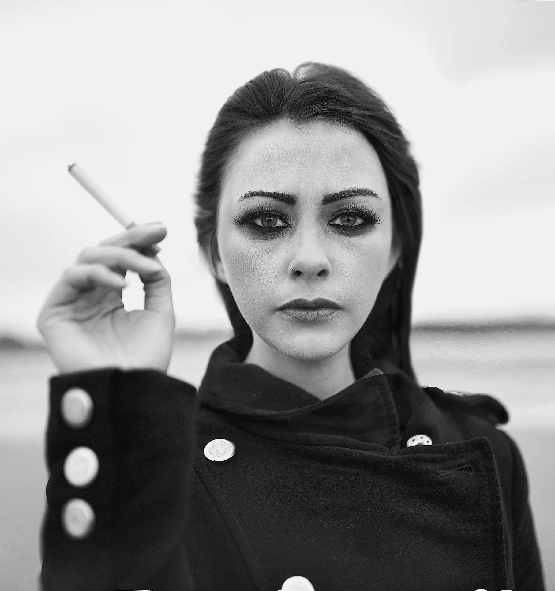 Black and white photo of a woman with dark hair, holding a cigarette, wearing a dark coat with metallic buttons, looking directly at the camera.