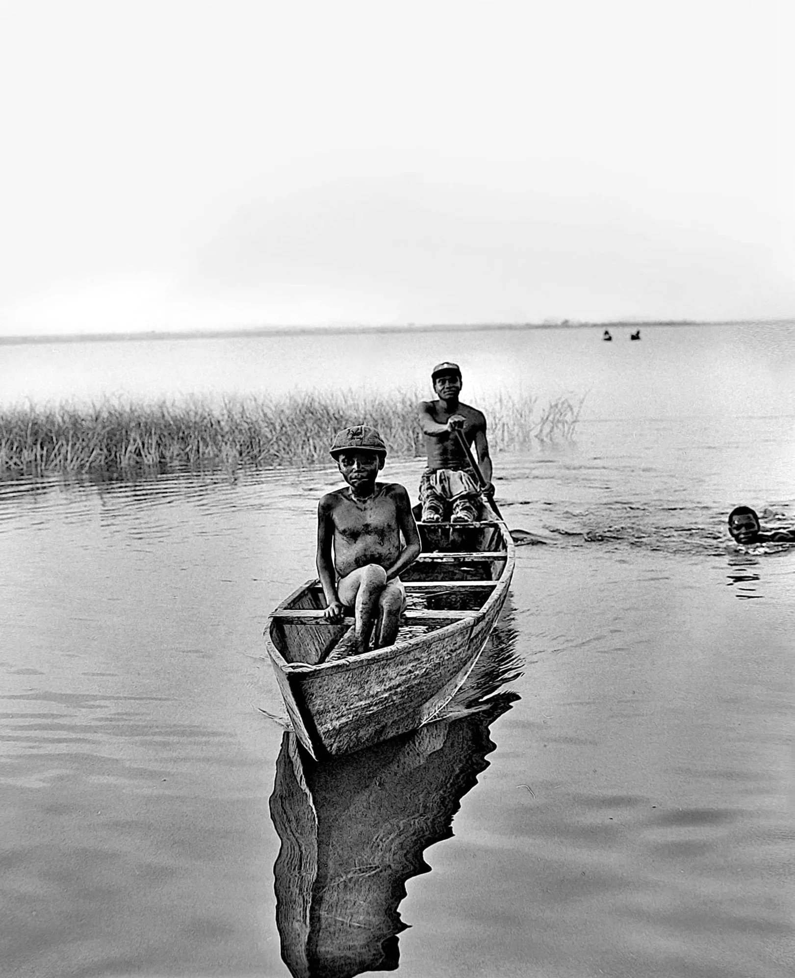 Black and white photo of three men in a small boat on a body of water, with one man holding a paddle and the other two sitting. In the background, there are two small boats on the water, tall grass along the shoreline, and a cloudy sky.