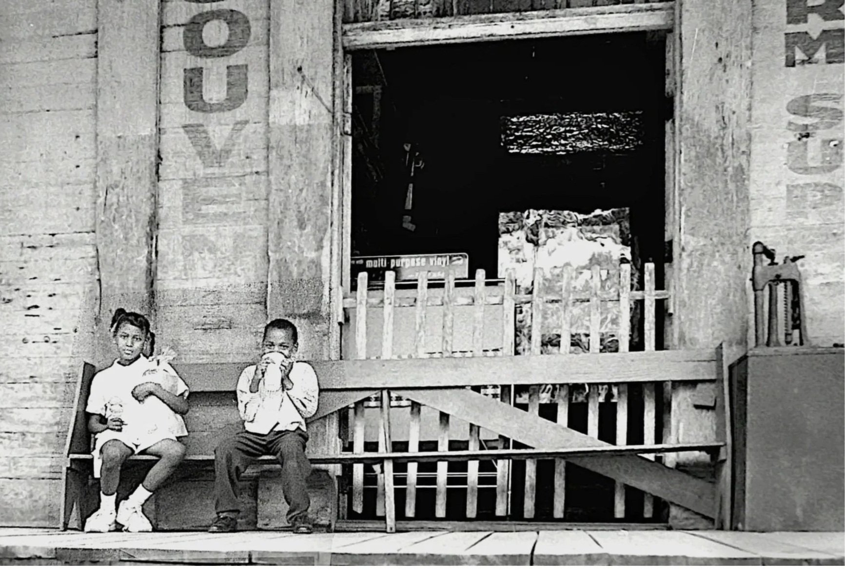 Black and white photo of two children sitting on a wooden porch in front of a rustic wooden building. The girl on the left is holding an ice cream cone, and the boy on the right is drinking from a cup. The background includes a staircase, wooden railings, and a doorway leading into the building.