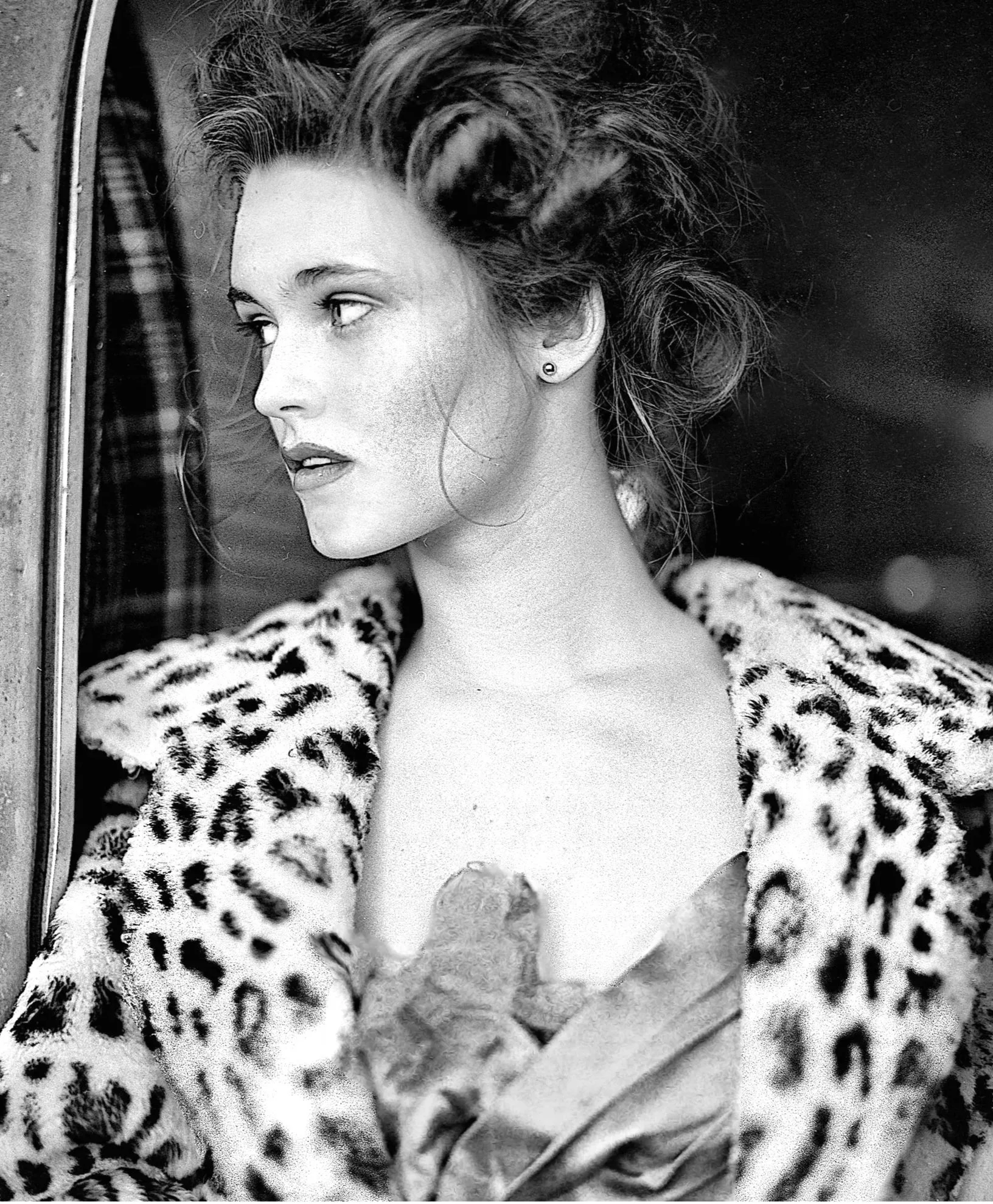 Black and white photo of a woman looking through a mirror, wearing leopard print clothing and earrings, with her hair styled in an updo.