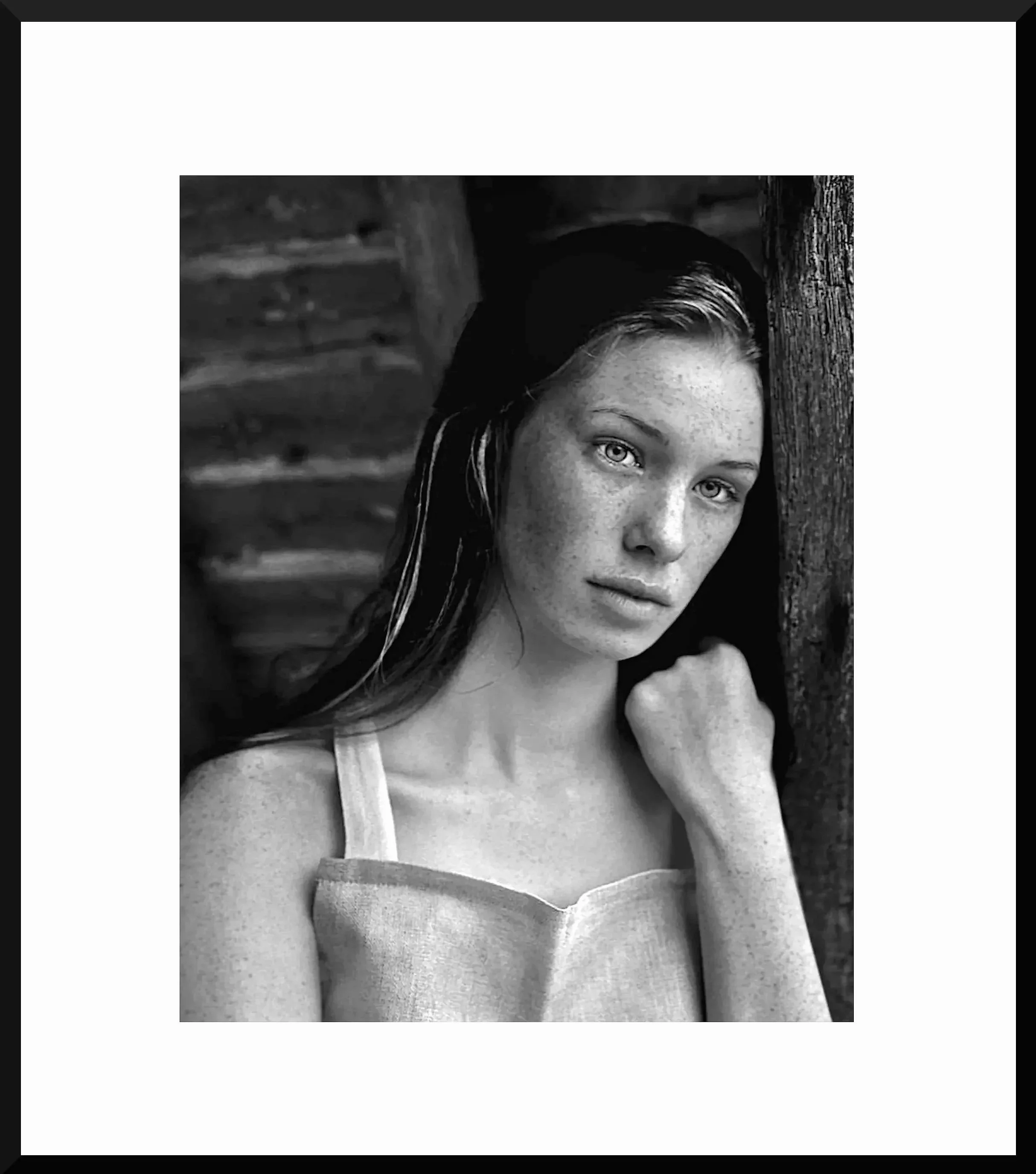 Black and white portrait of a young woman with light eyes, freckles, and long dark hair, standing next to a wooden post.
