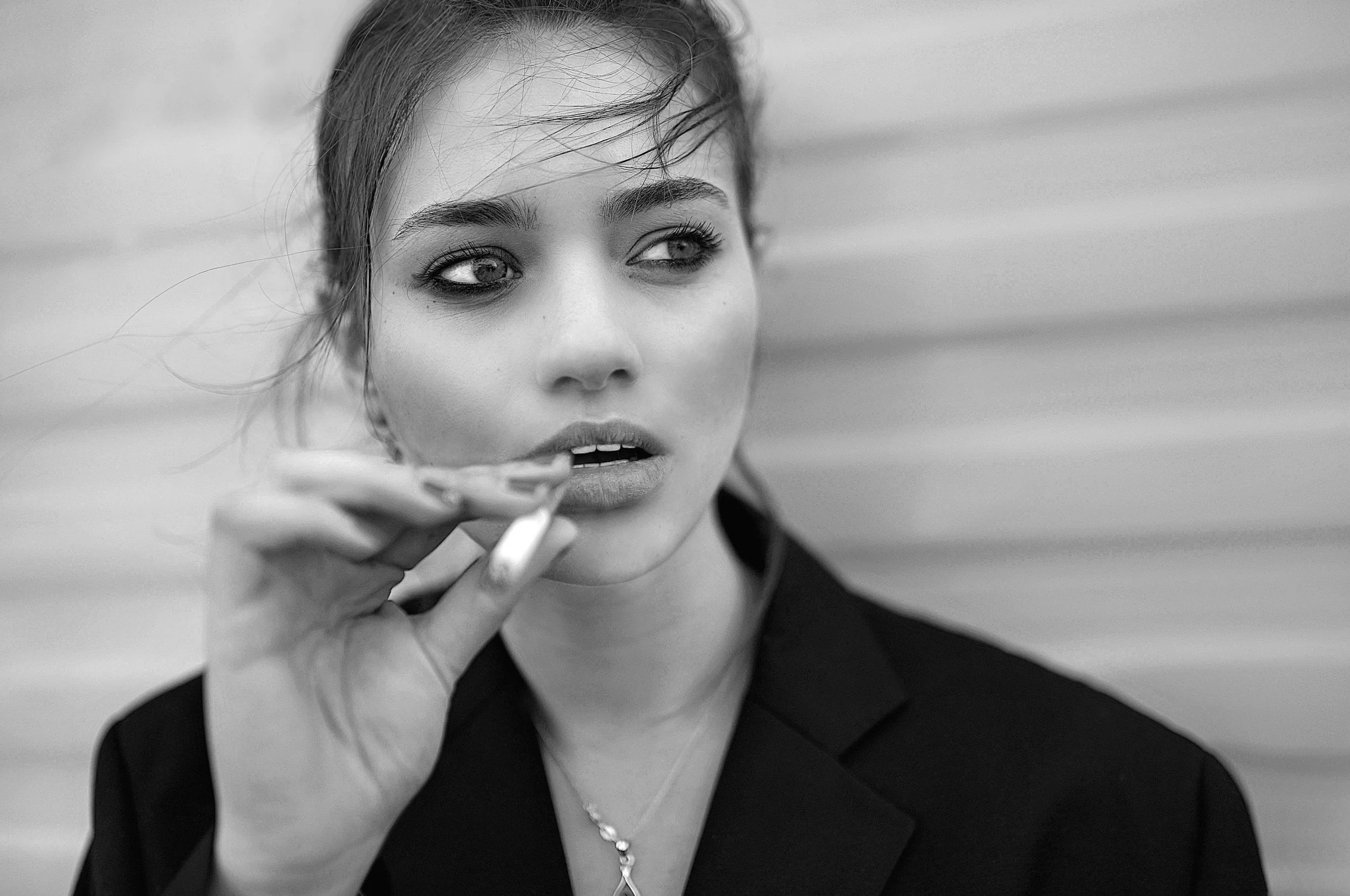 A young woman with light skin, dark eye makeup, and wet hair, wearing a black blazer and a necklace, holding a cigarette near her lips, standing against a horizontal striped background.