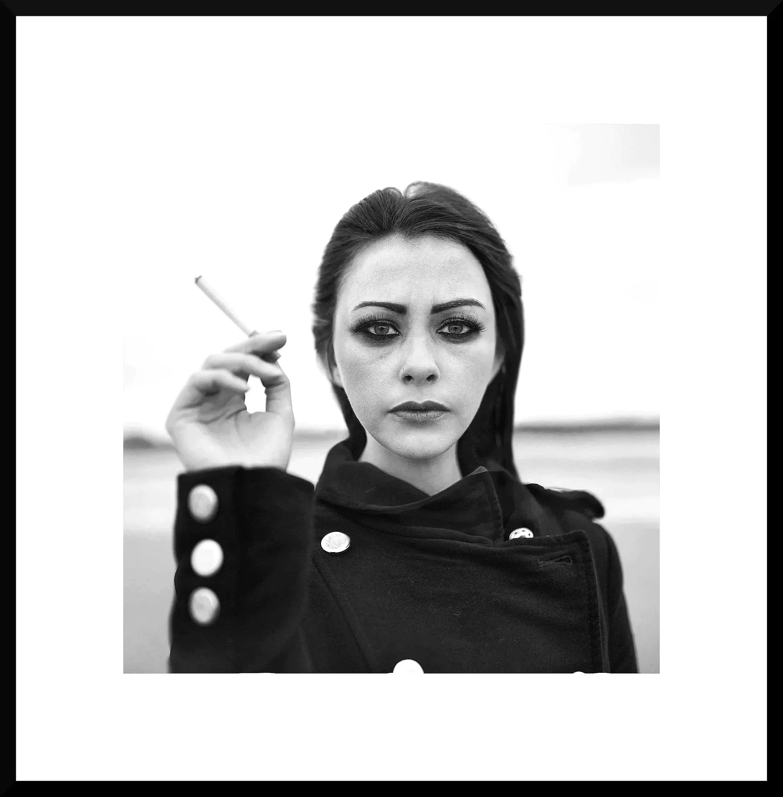 A black and white photo of a woman with dark hair and makeup, wearing a dark jacket with buttons, holding a cigarette in her right hand and staring directly at the camera.