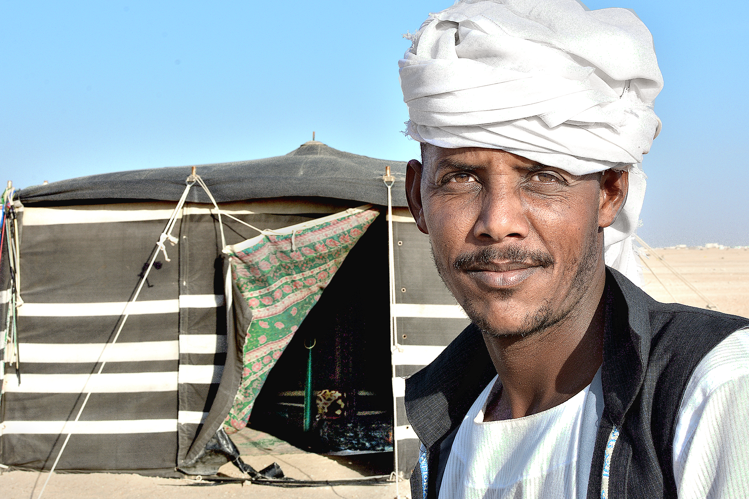 A man wearing a white turban and black vest stands outdoors in front of a striped tent with a blue sky background.