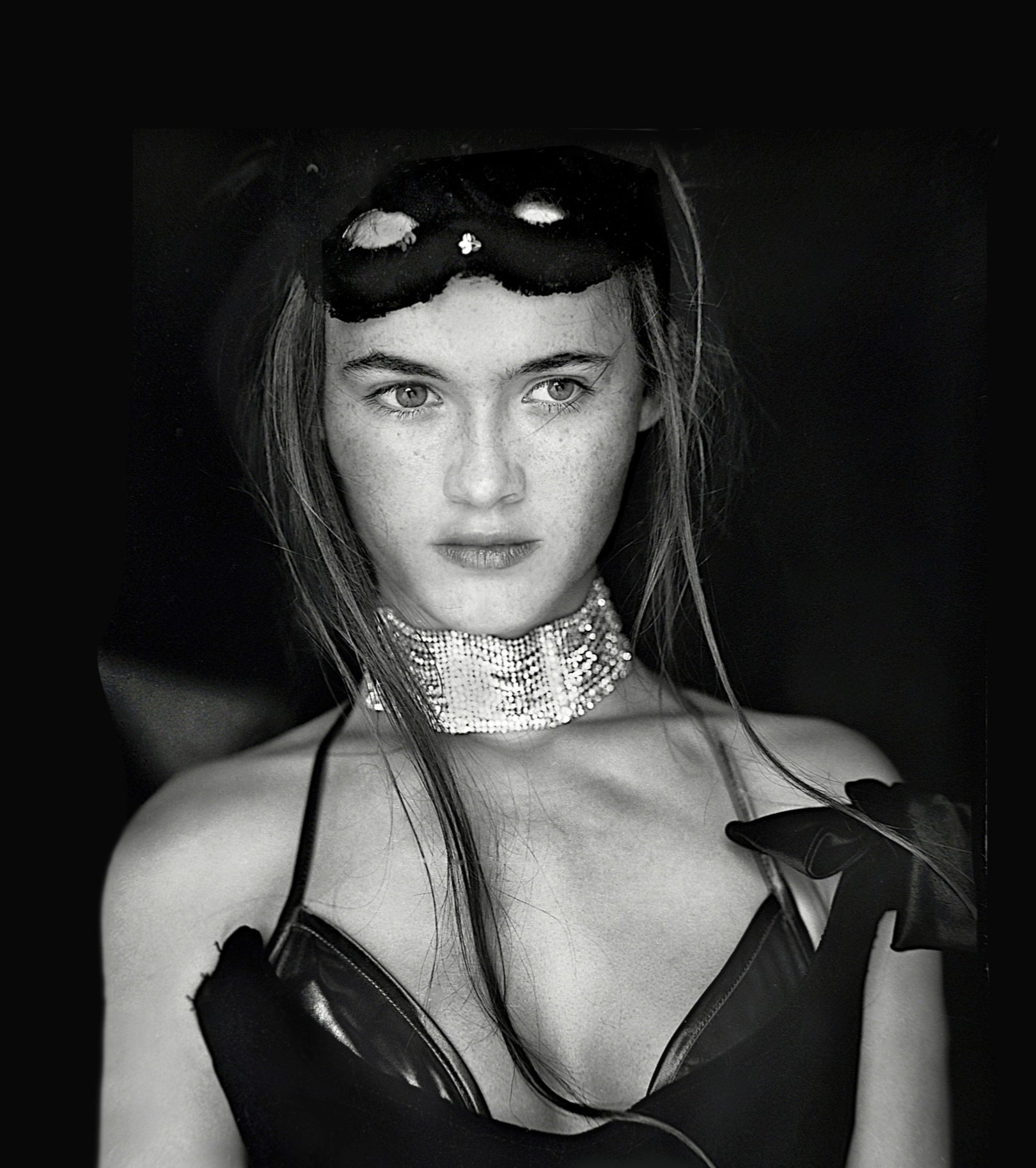 Black and white portrait of a young woman wearing a shiny, strappy top, gloves, and a sparkling choker necklace. She has light-colored eyes, freckles, and is holding her shoulders with gloved hands. She wears a black mask on her head and gazes directly at the camera.
