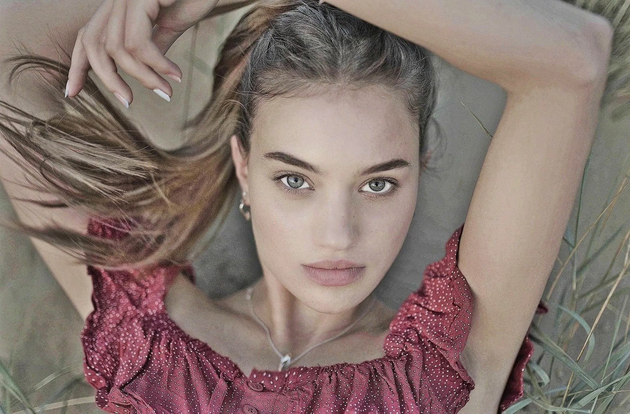 A young woman with blue eyes and long blonde hair lying on the ground, wearing a red polka-dot top and silver jewelry, looking directly at the camera with her arms raised above her head.