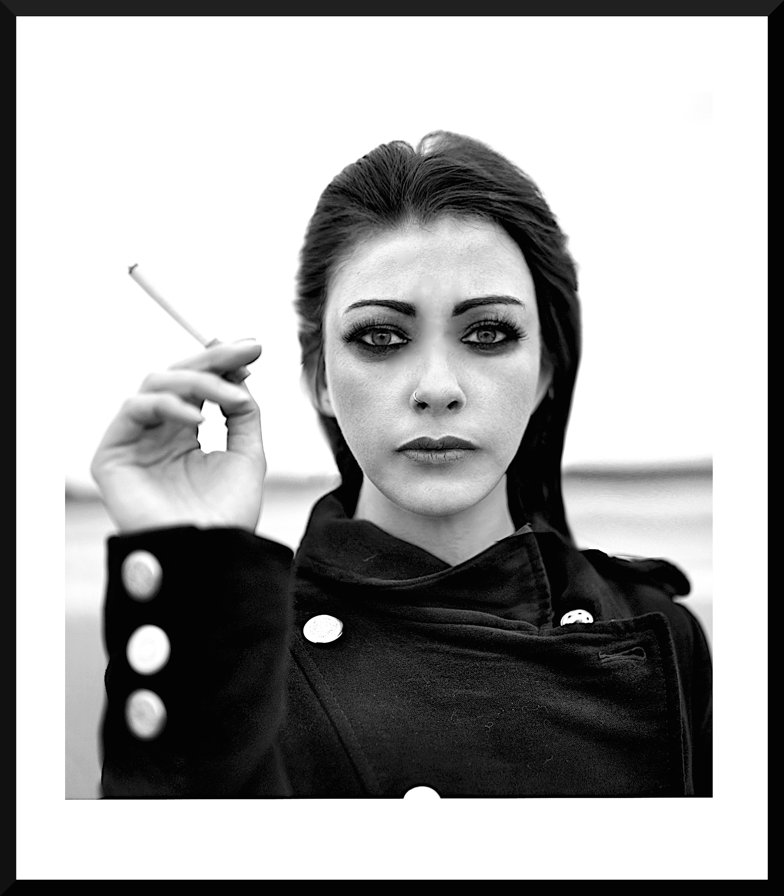A black and white photo of a woman with dark hair and makeup, holding a cigarette in her right hand, wearing a dark coat with large buttons.