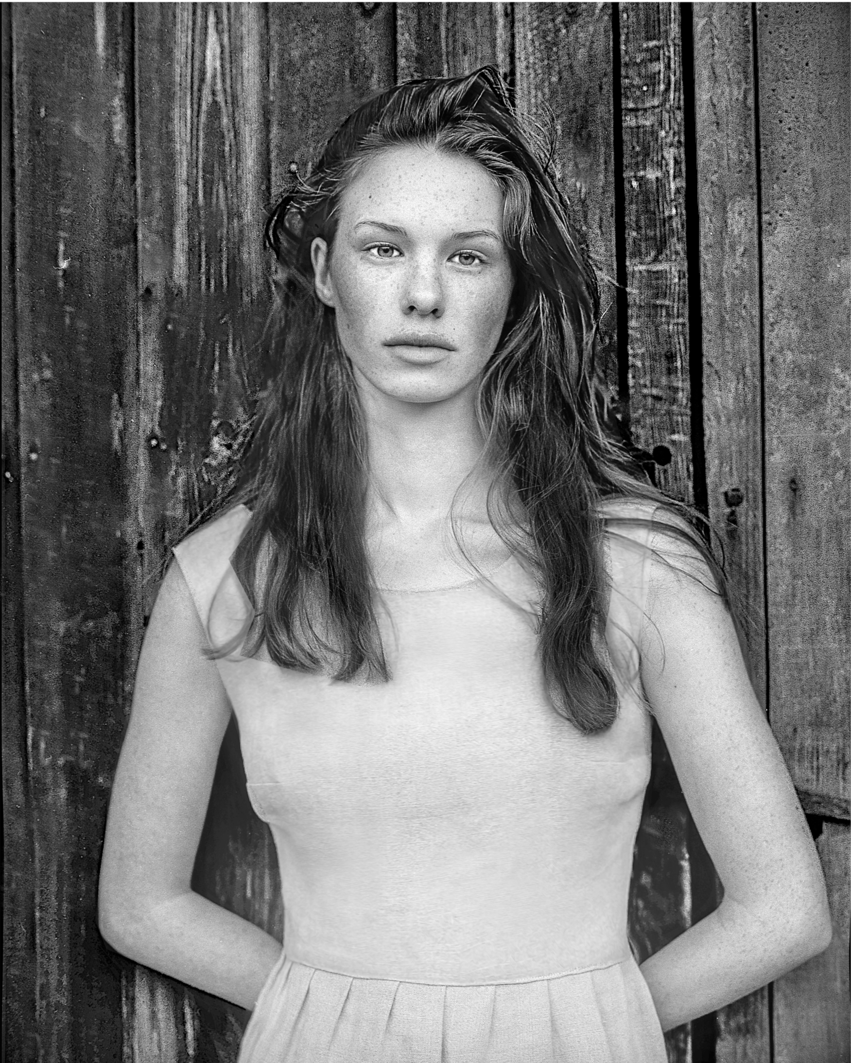 Black and white photo of a young woman with long, wavy hair standing in front of a rustic wooden fence. She is wearing a light-colored sleeveless top and a pleated skirt, with her left arm resting on the fence behind her.