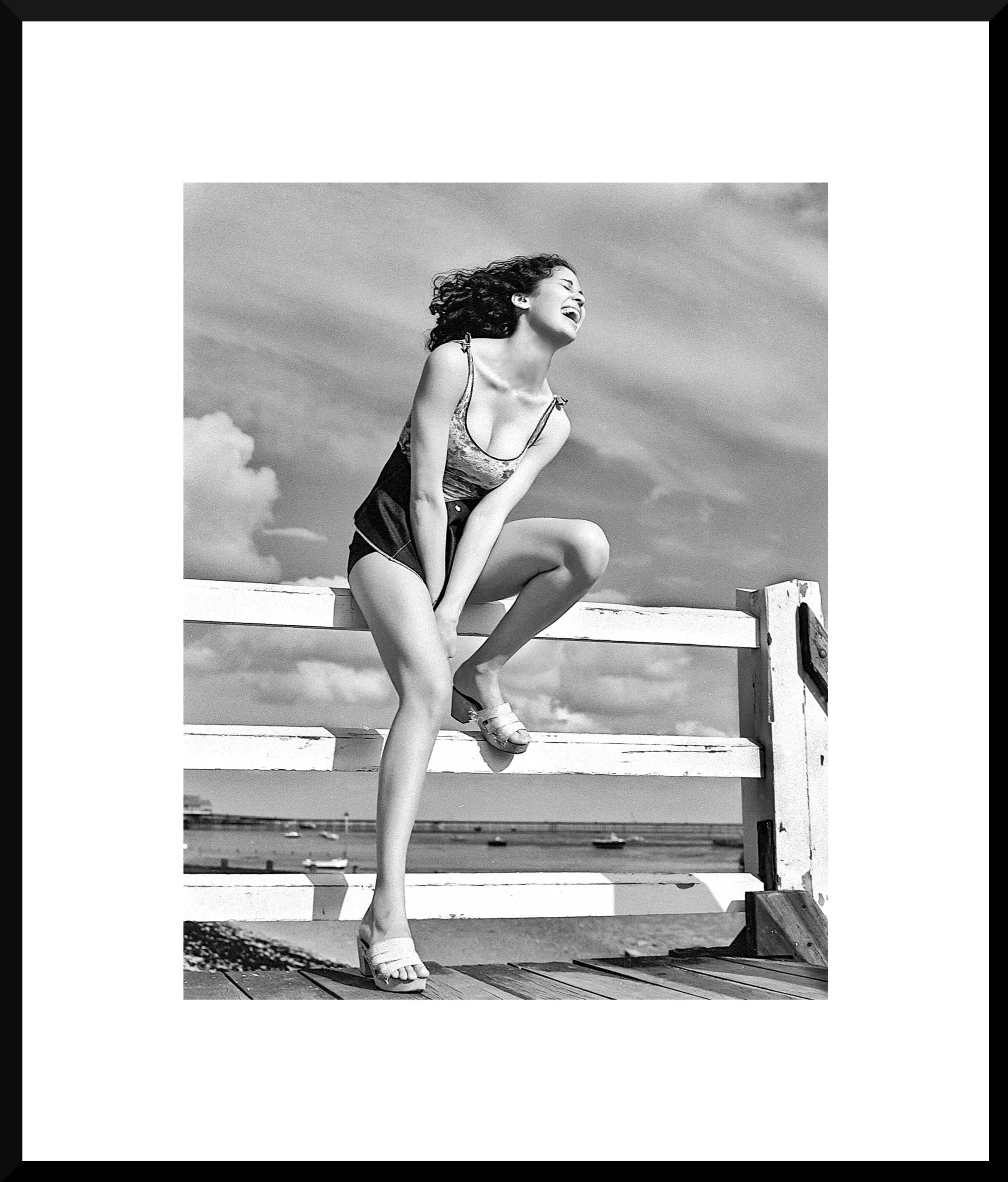 A woman laughing, crouching on a wooden pier next to a white railing with water and boats in the background, in black and white.