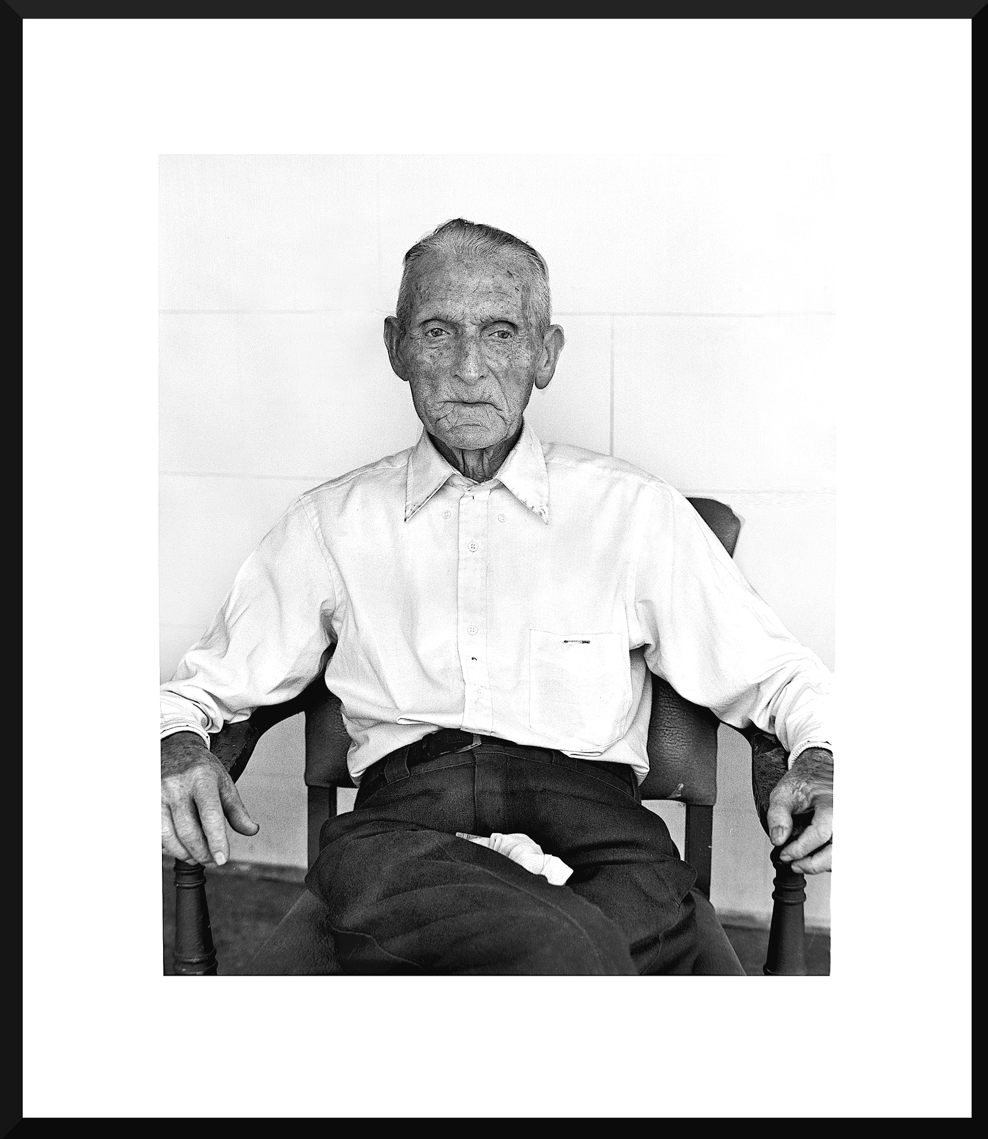 A black-and-white photo of an elderly man sitting in a chair against a plain wall, wearing a button-up shirt and pants.