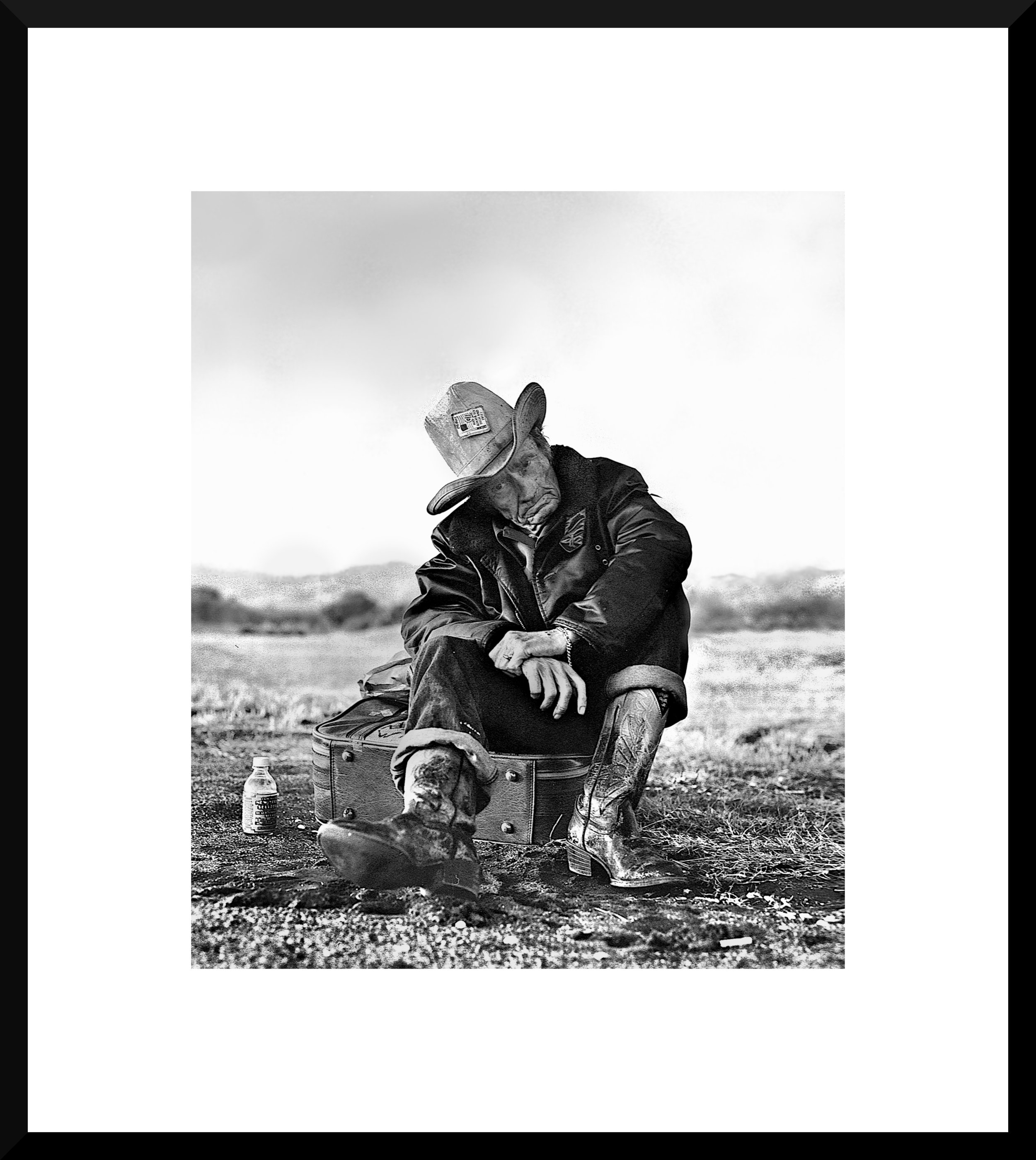 A black and white photograph of an elderly man sitting on a suitcase outdoors, wearing a cowboy hat, leather jacket, and cowboy boots, with a small dog in front of him and a water bottle on the ground nearby.