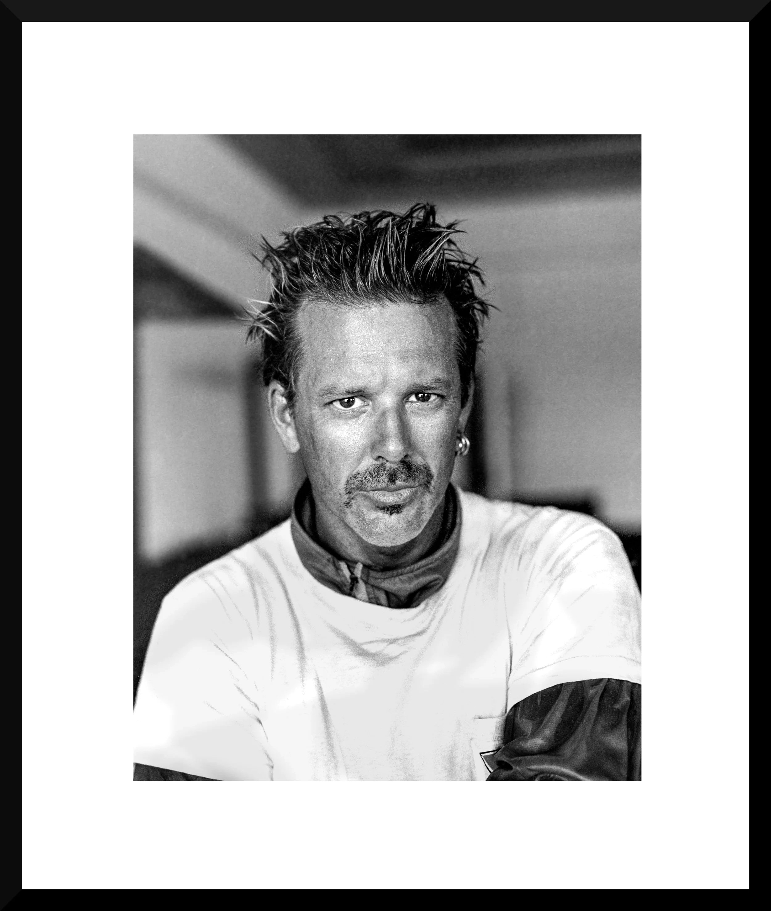 Black and white portrait of a man with styled spiky hair, a mustache, and an earring, wearing a white shirt and looking directly at the camera.