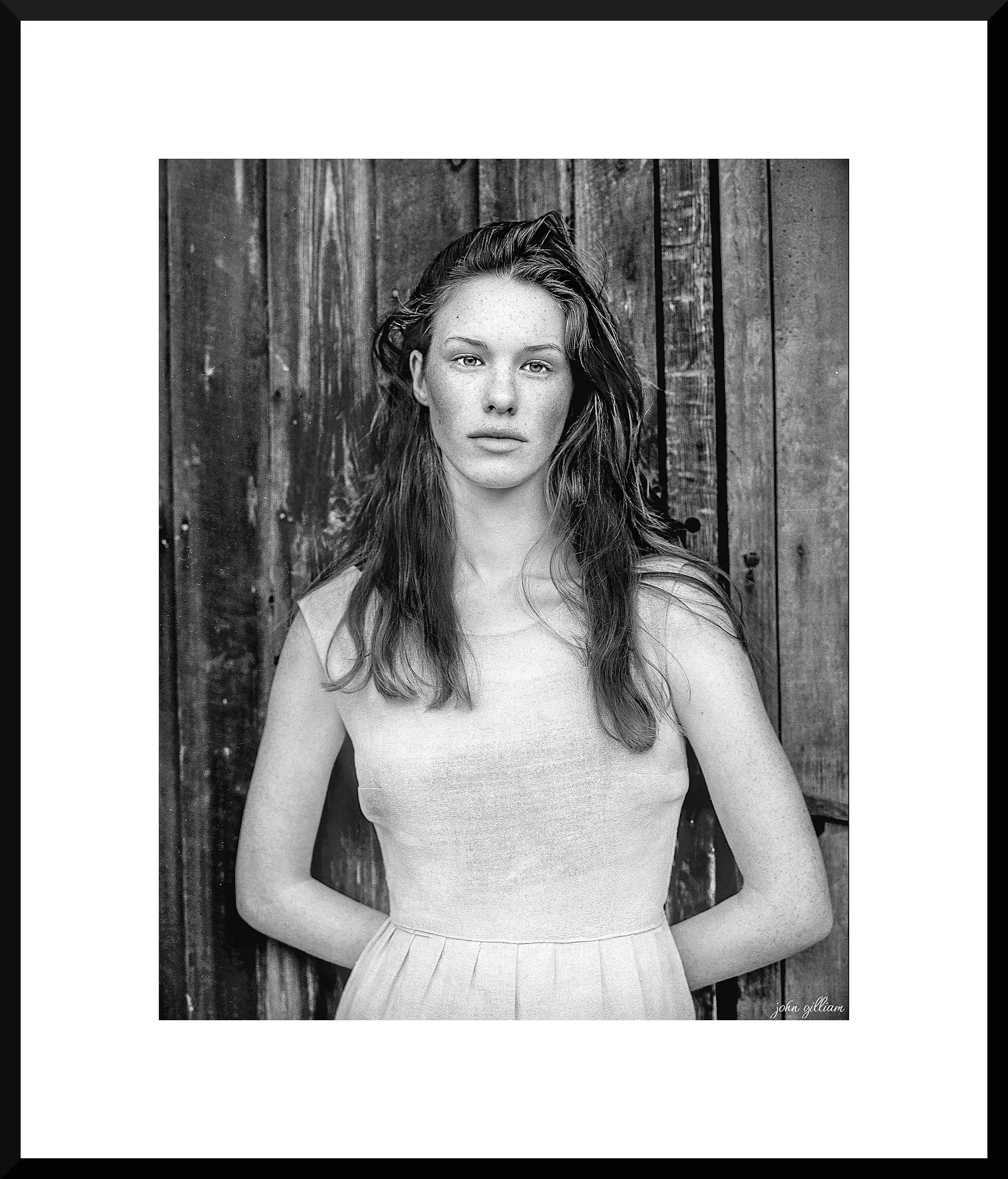 Black and white portrait of a young woman with long wavy hair standing in front of a wooden backdrop.