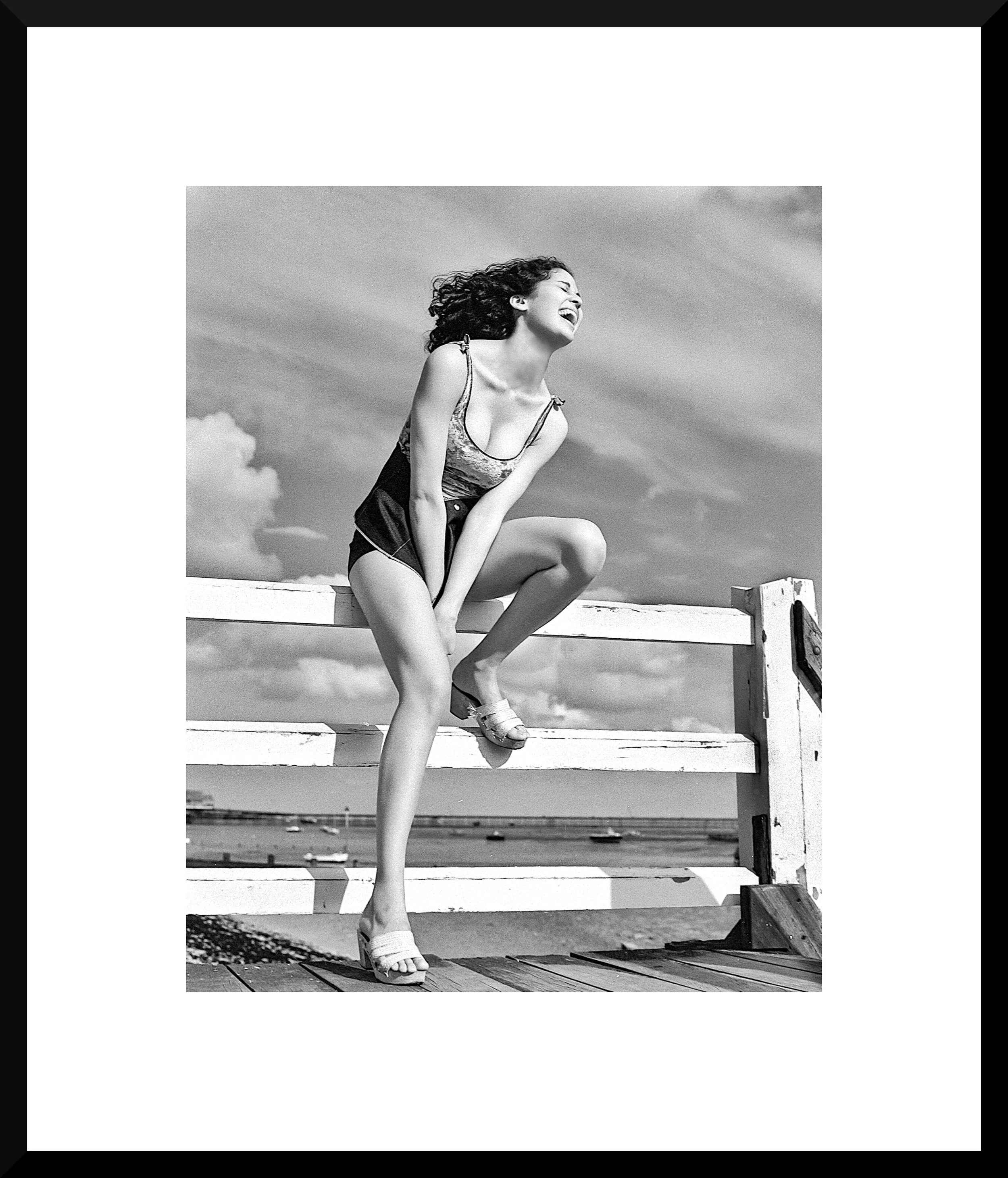 A woman laughing and posing on a seaside pier in black and white, with sky and clouds in the background.