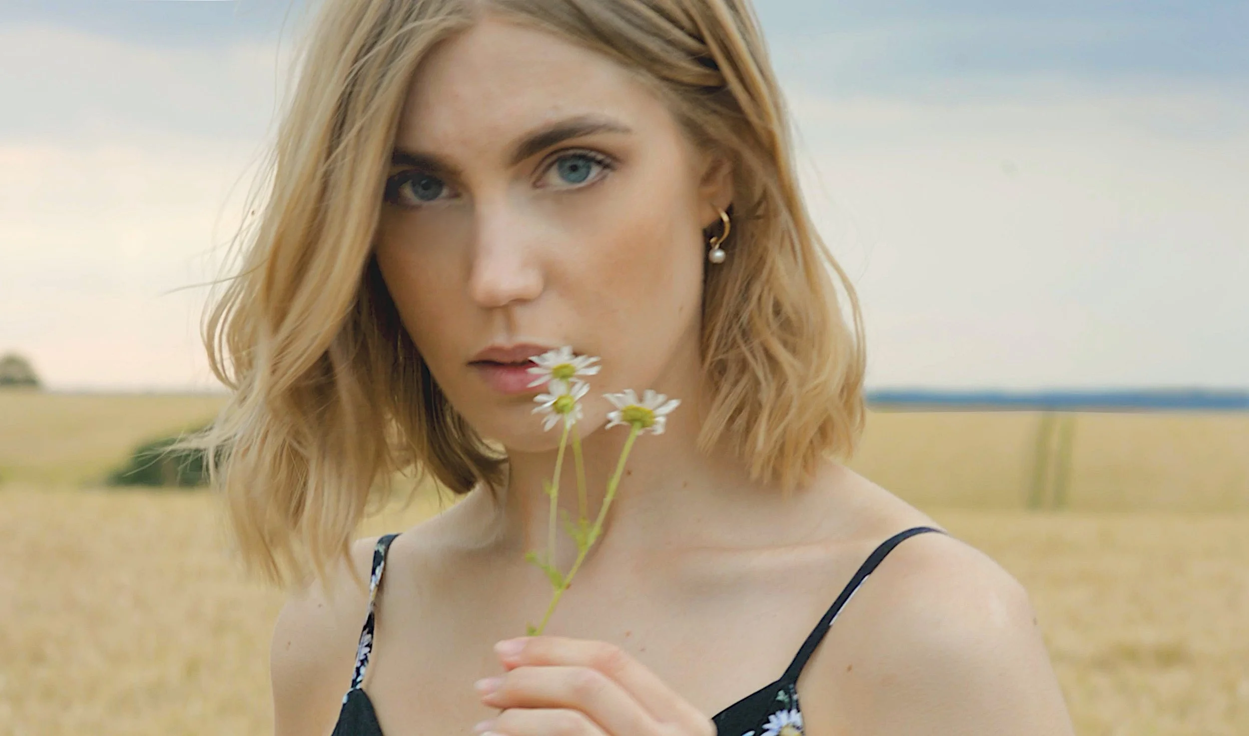 A woman with shoulder-length blonde hair and blue eyes holding a small bunch of white daisies near her lips in an open field with a cloudy sky in the background.