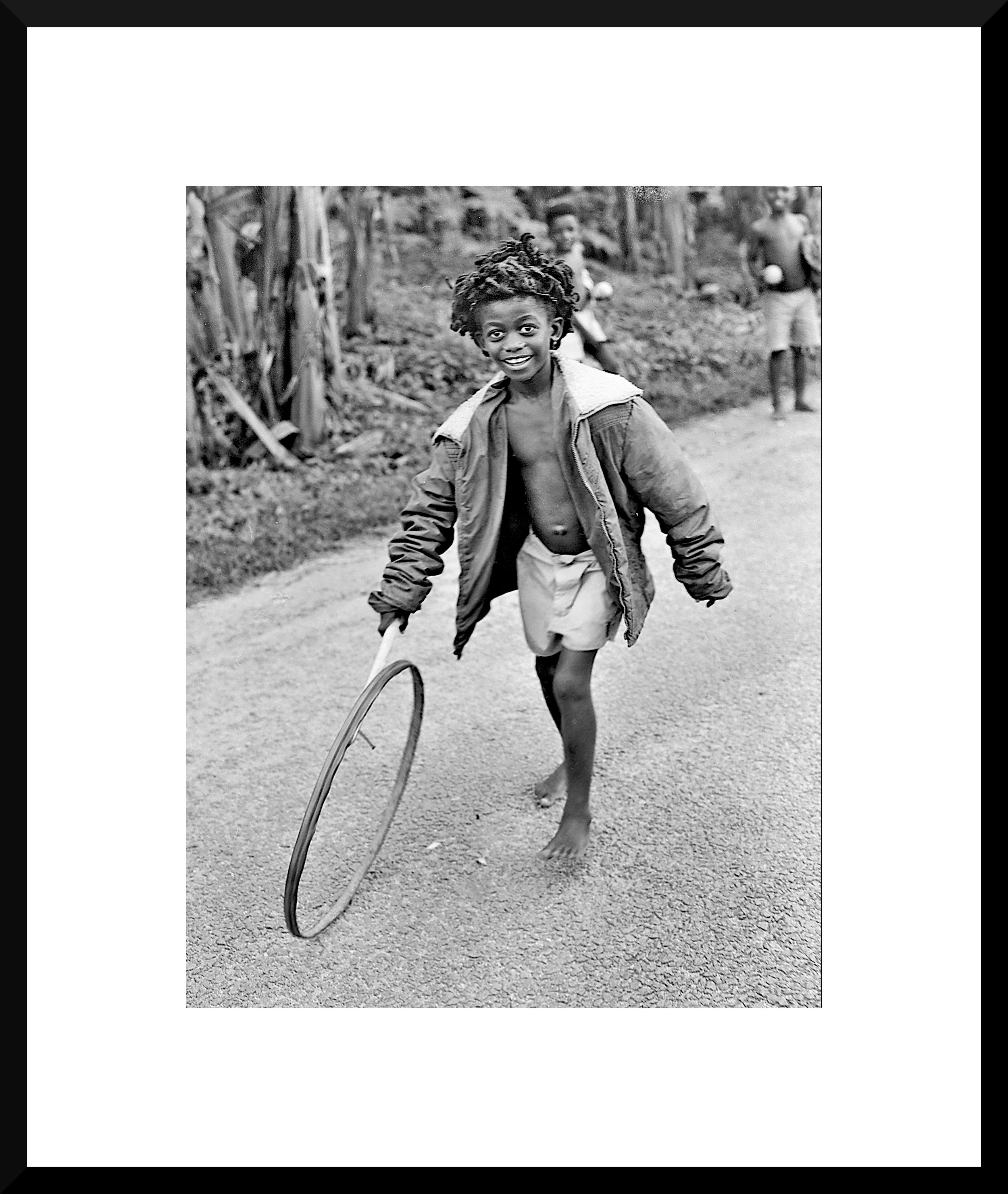 A smiling young boy walking barefoot on a dirt road, holding a hula hoop, wearing a jacket and shorts, with other children in the background.