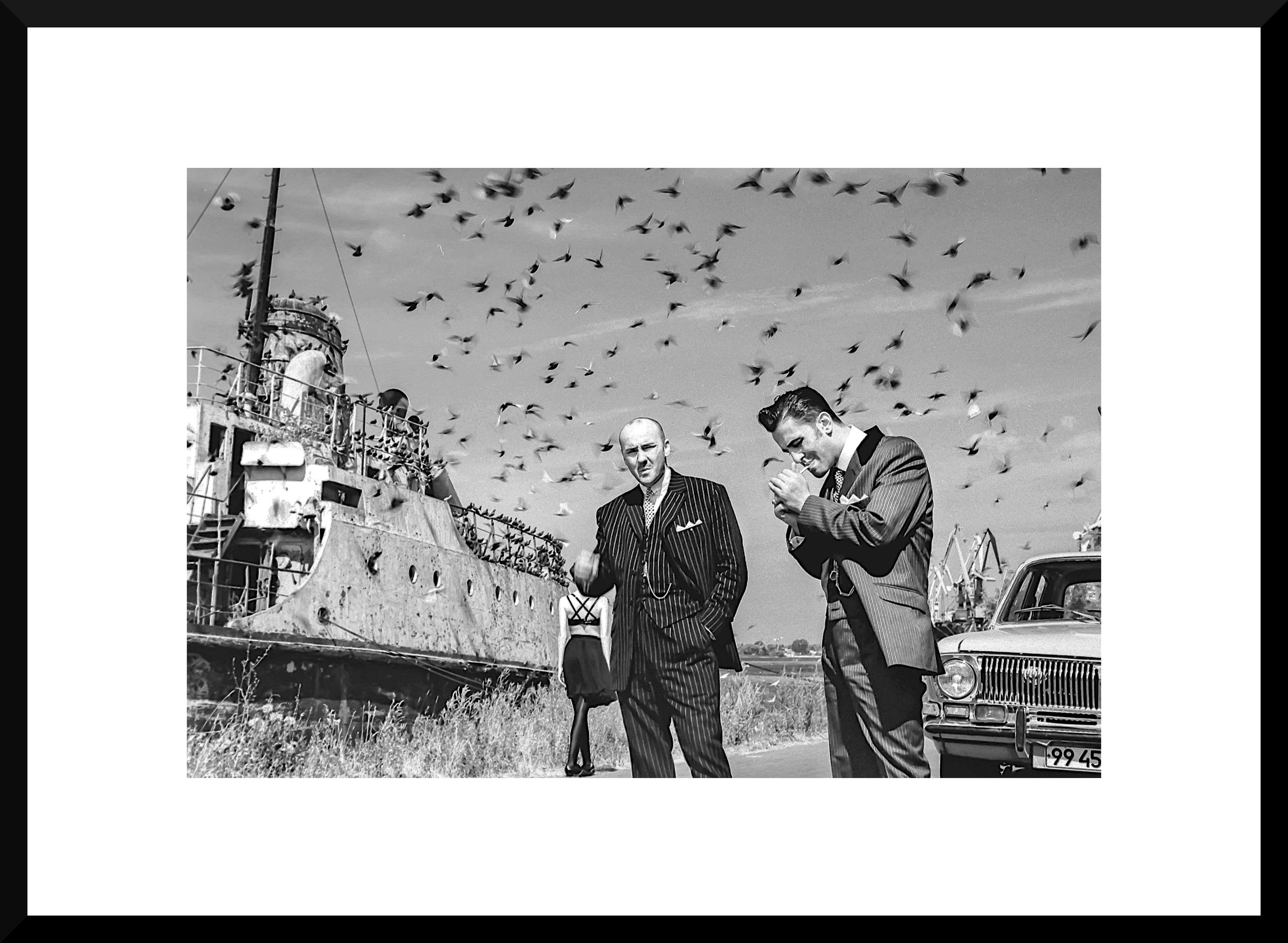 Black and white photo of two men in suits standing near an old cruise ship, with pigeons flying overhead, an older car in the background, and a woman walking away in the distance.
