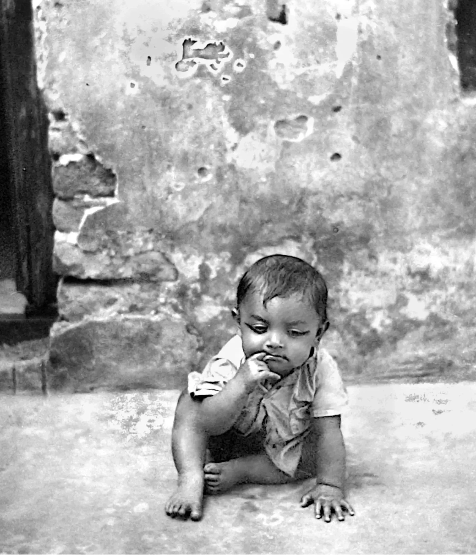 A young boy is crouched on the ground with his fingers in his mouth, in front of a rough stone wall. The photograph is in black and white.