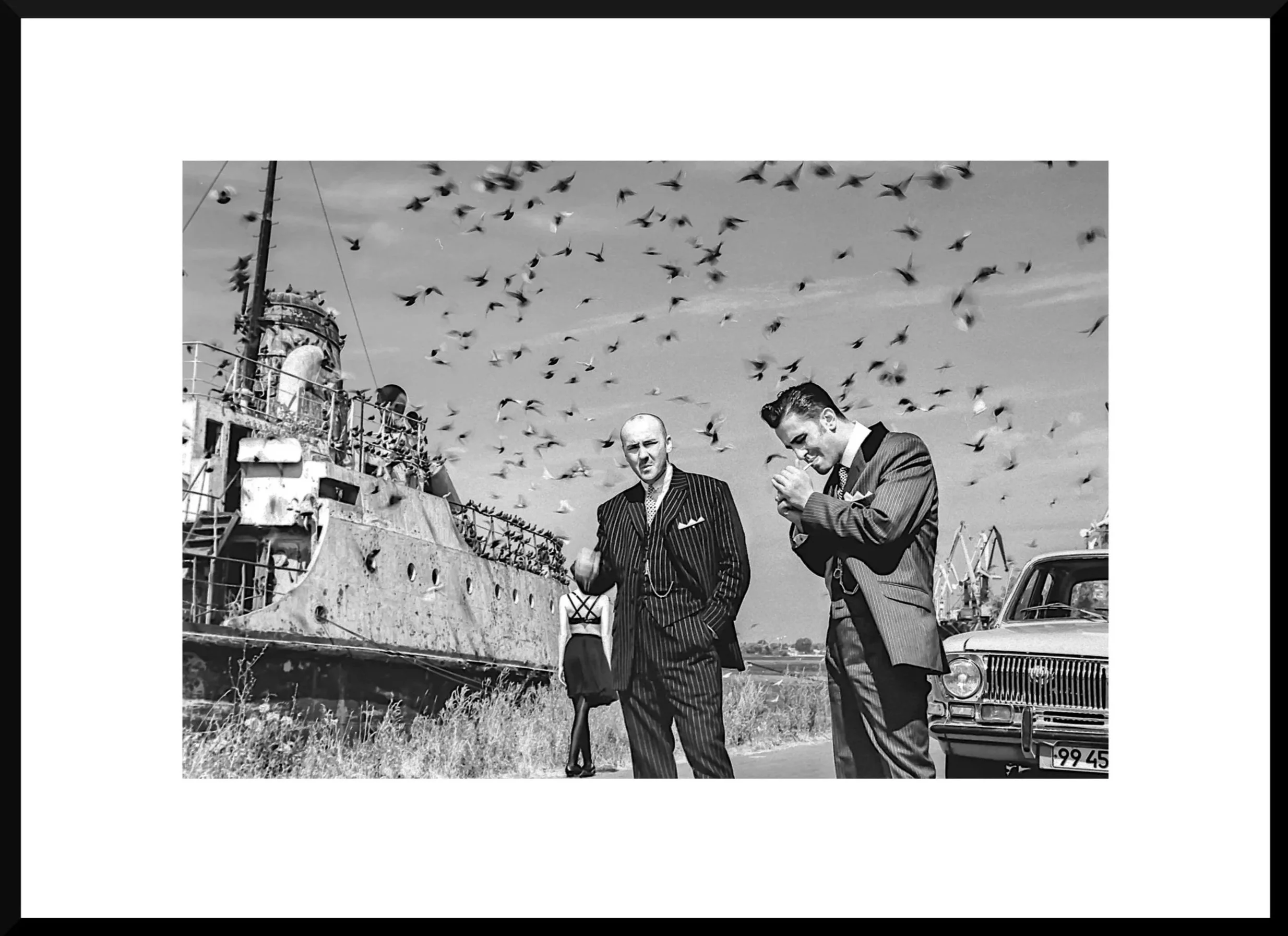Black and white photo of two men in suits standing near an old car and a rusty shipwreck, with a woman in the background and birds flying overhead.