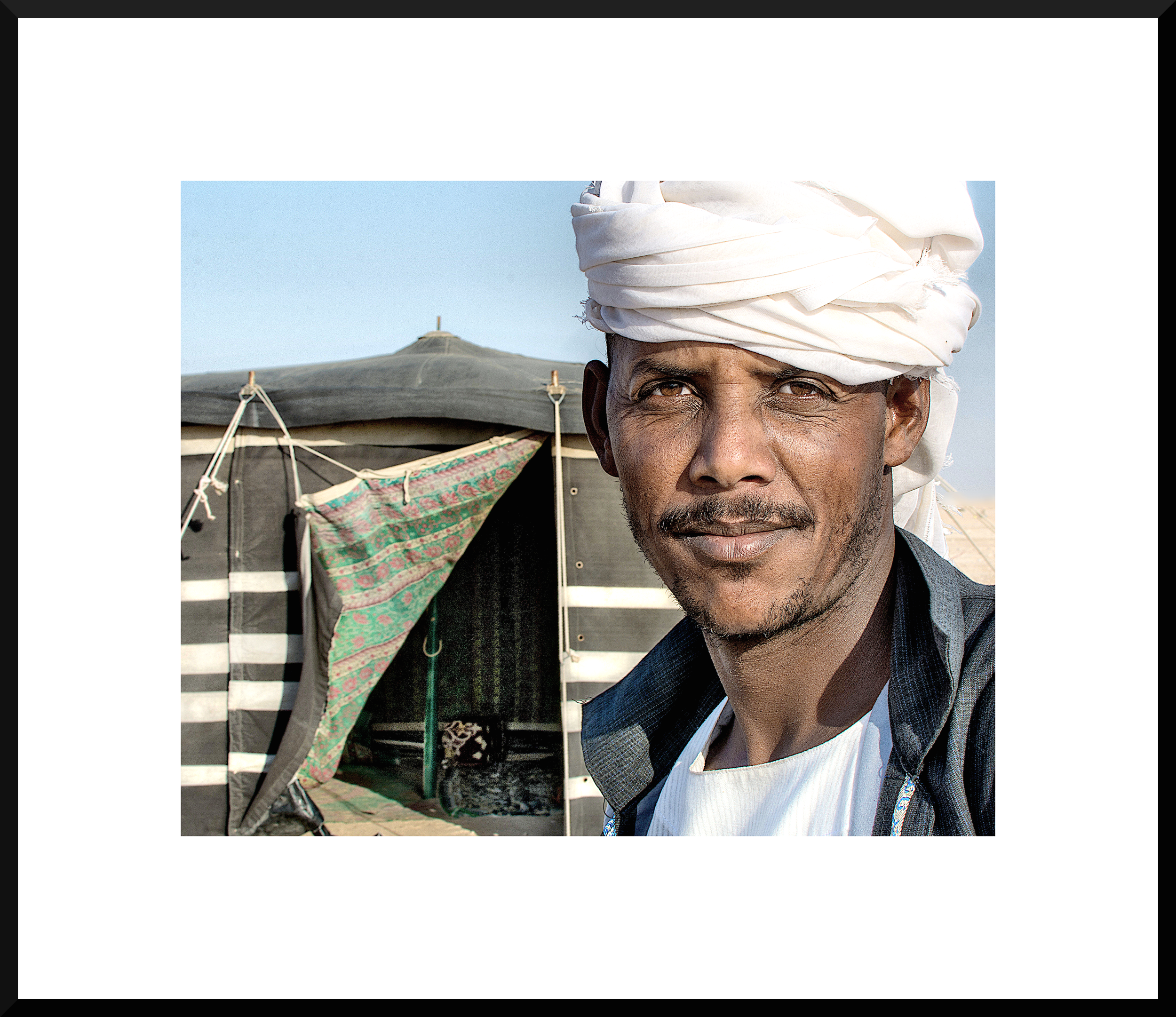 Close-up of a man wearing a white turban, standing in front of a striped tent with an open entrance, in a desert landscape.
