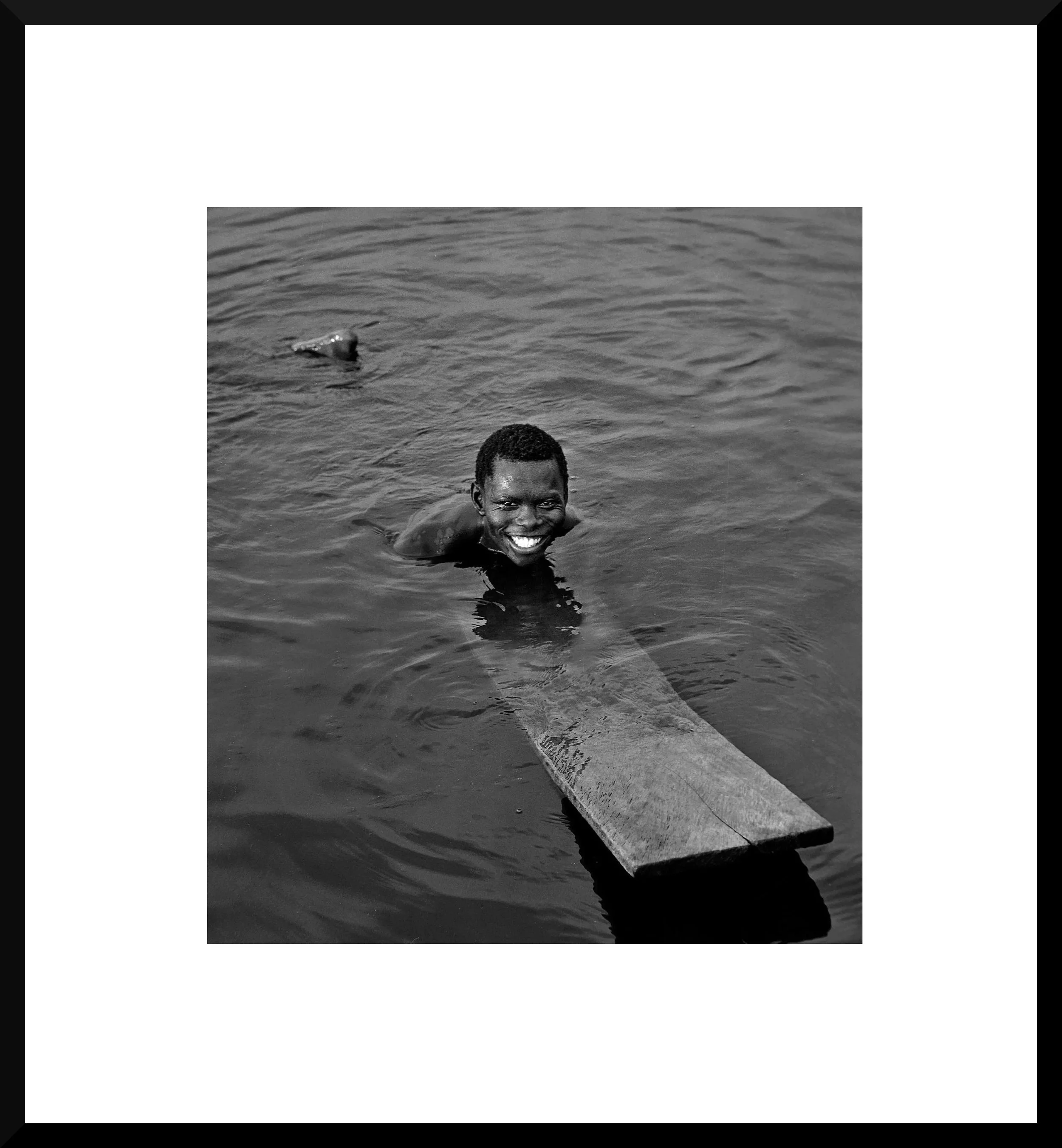 A smiling boy swimming in water, resting on a wooden board, in a black-and-white photo.