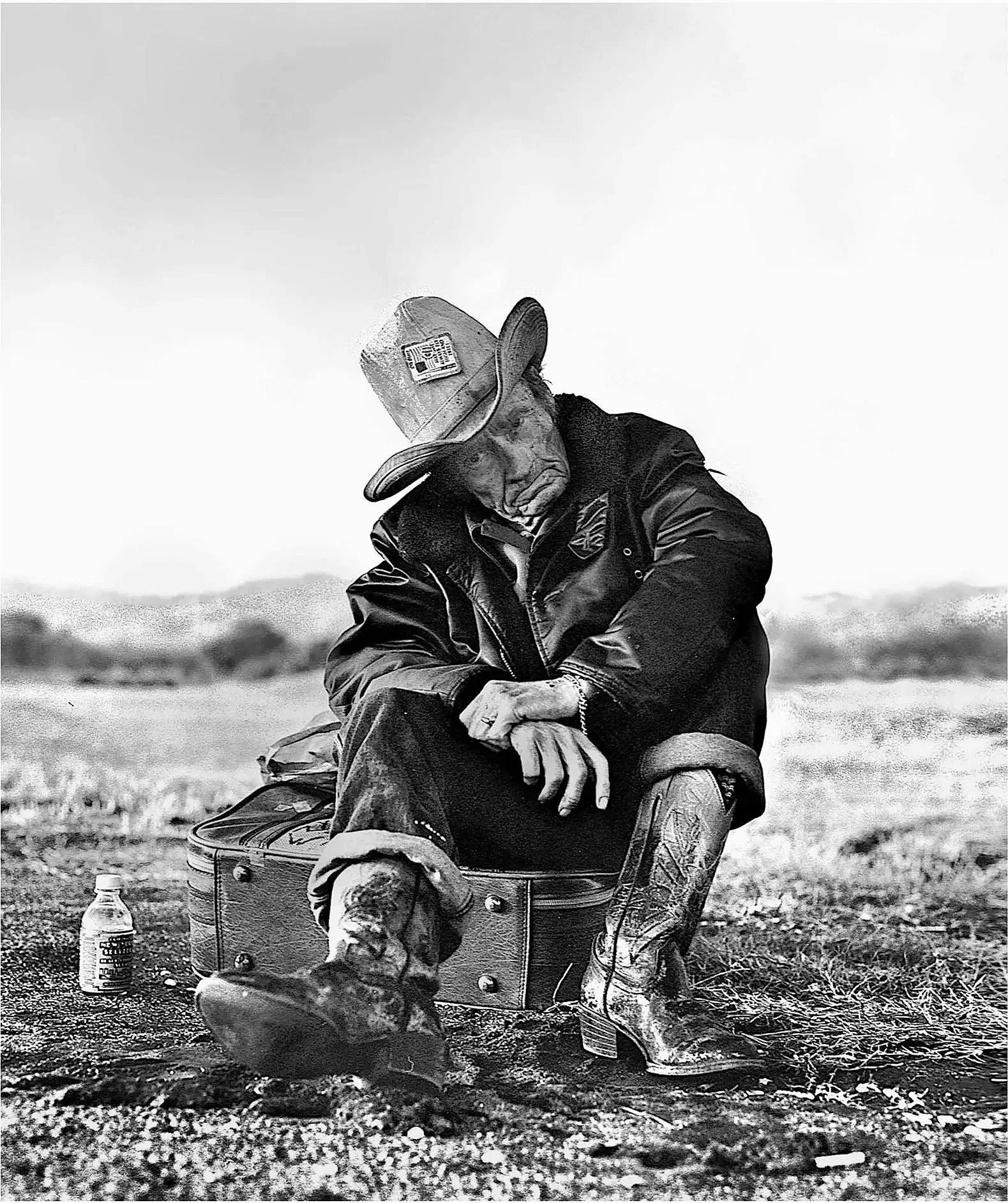 A black and white photo of a man sitting on a portable seat outdoors. He is wearing a cowboy hat, sunglasses, a jacket, and jeans. There is a water bottle and a small cooler beside him on the ground.