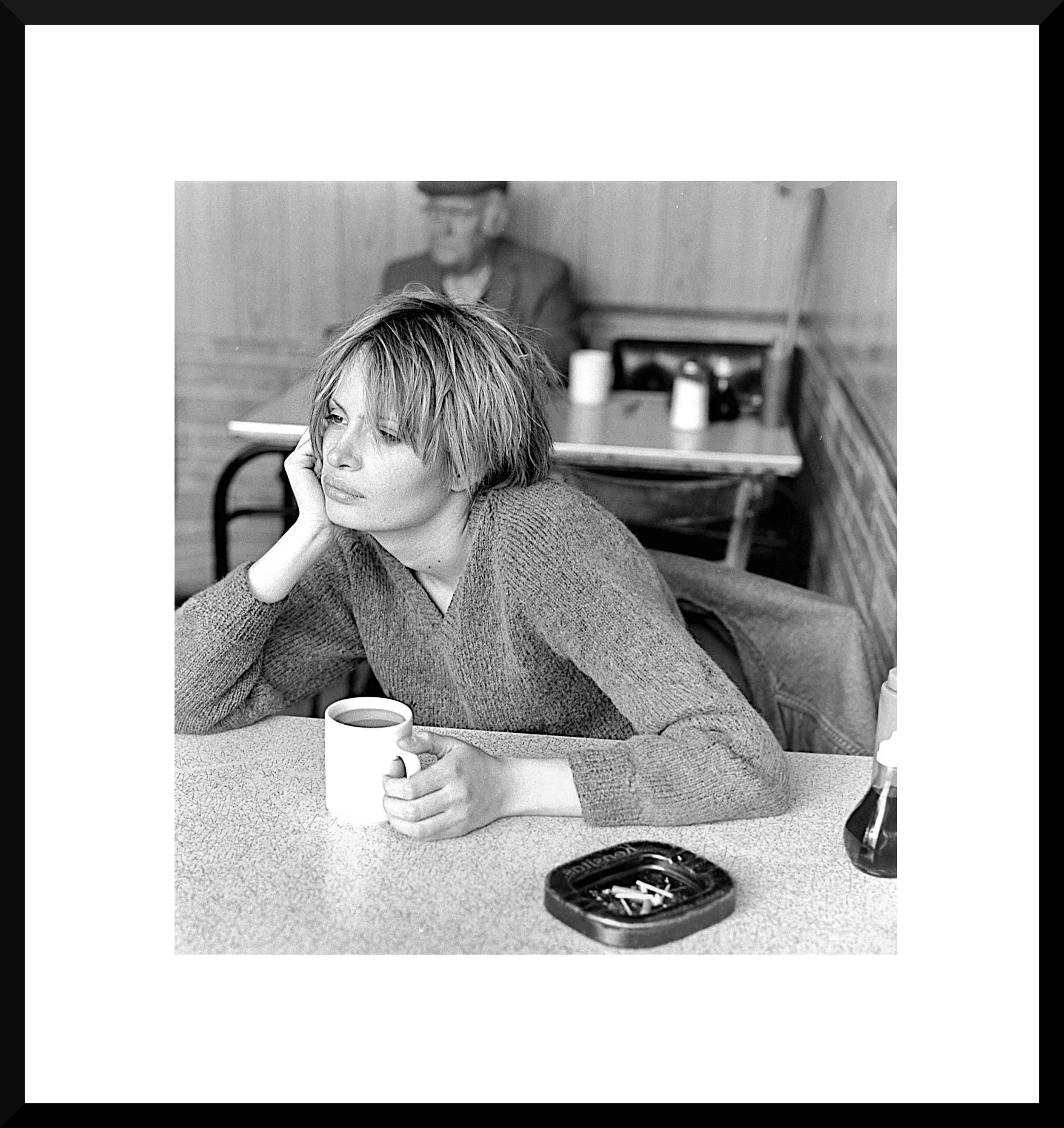 A woman with short hair sitting at a table in a cafe, holding a mug of coffee, with a thoughtful expression. There is an ashtray with cigarette butts on the table, and a man sitting in the background.