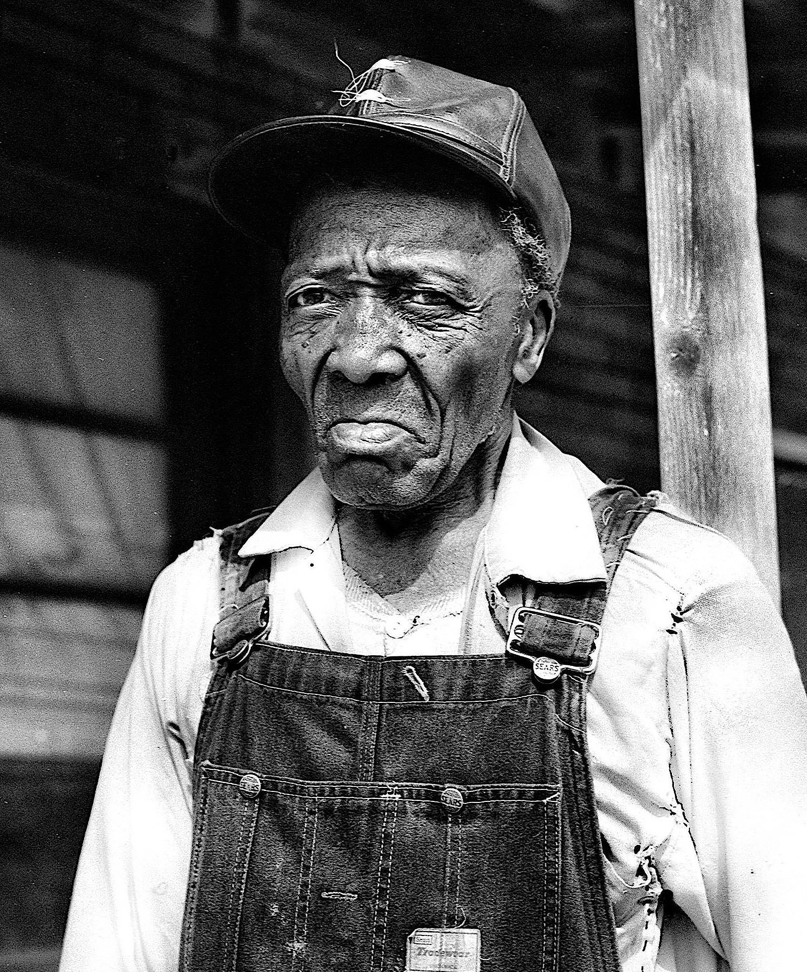 Black and white photograph of an elderly man wearing a cap, a collared shirt, and denim overalls, standing outdoors with a wooden post visible in the background.