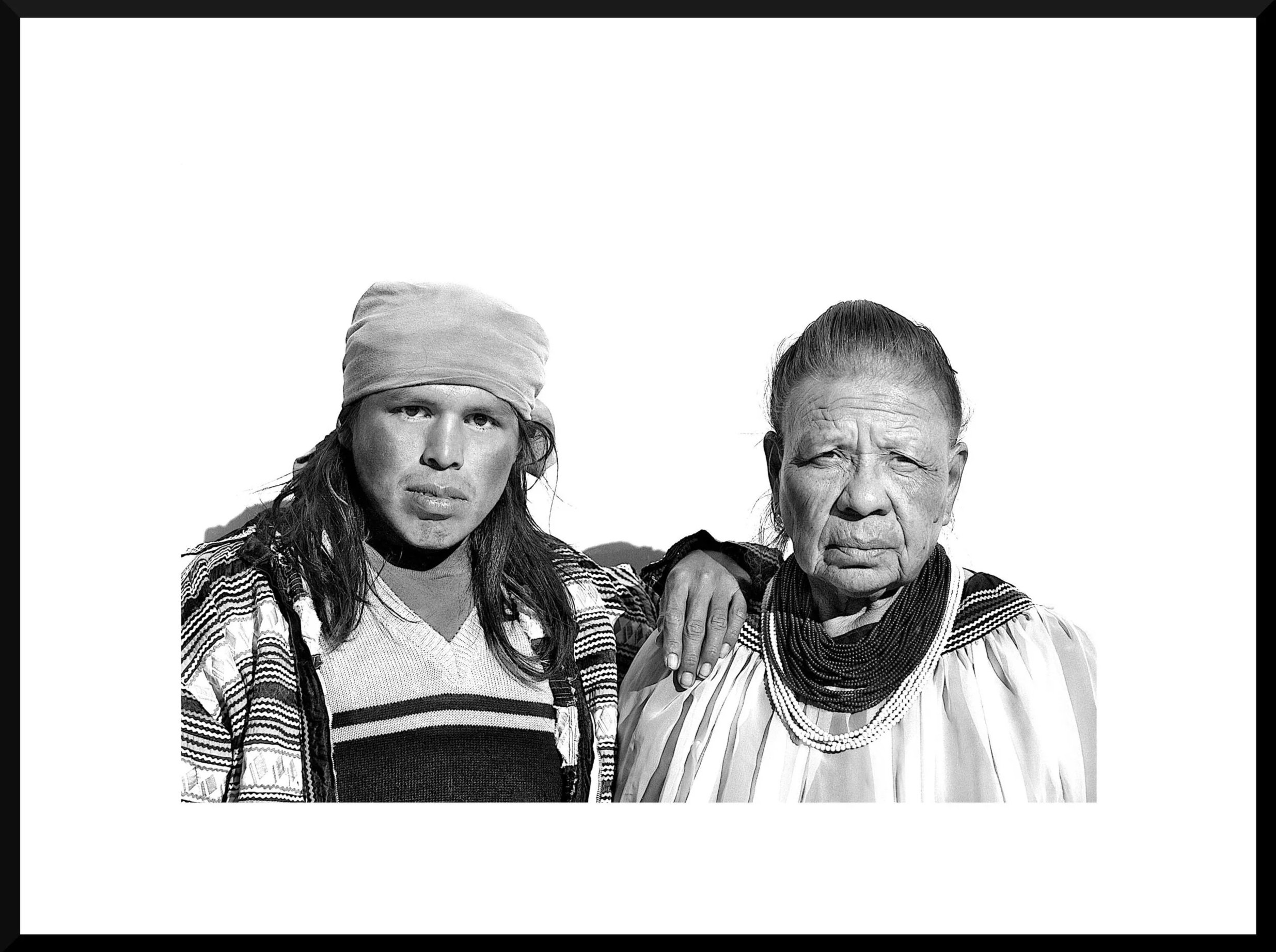 Black and white photograph of two indigenous women standing outdoors against a plain sky, with the woman on the left wearing a headscarf and traditional patterned clothing, and the woman on the right wearing a blouse, scarf, and layered necklaces.