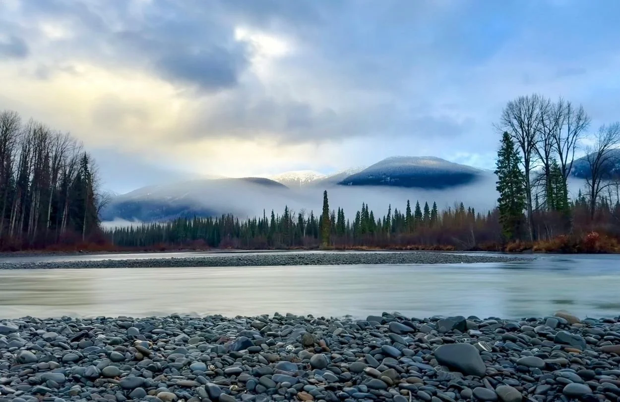 relationship therapy across BC and A river with a rocky shore in the foreground, surrounded by evergreen trees, with mountains and clouds in the background.