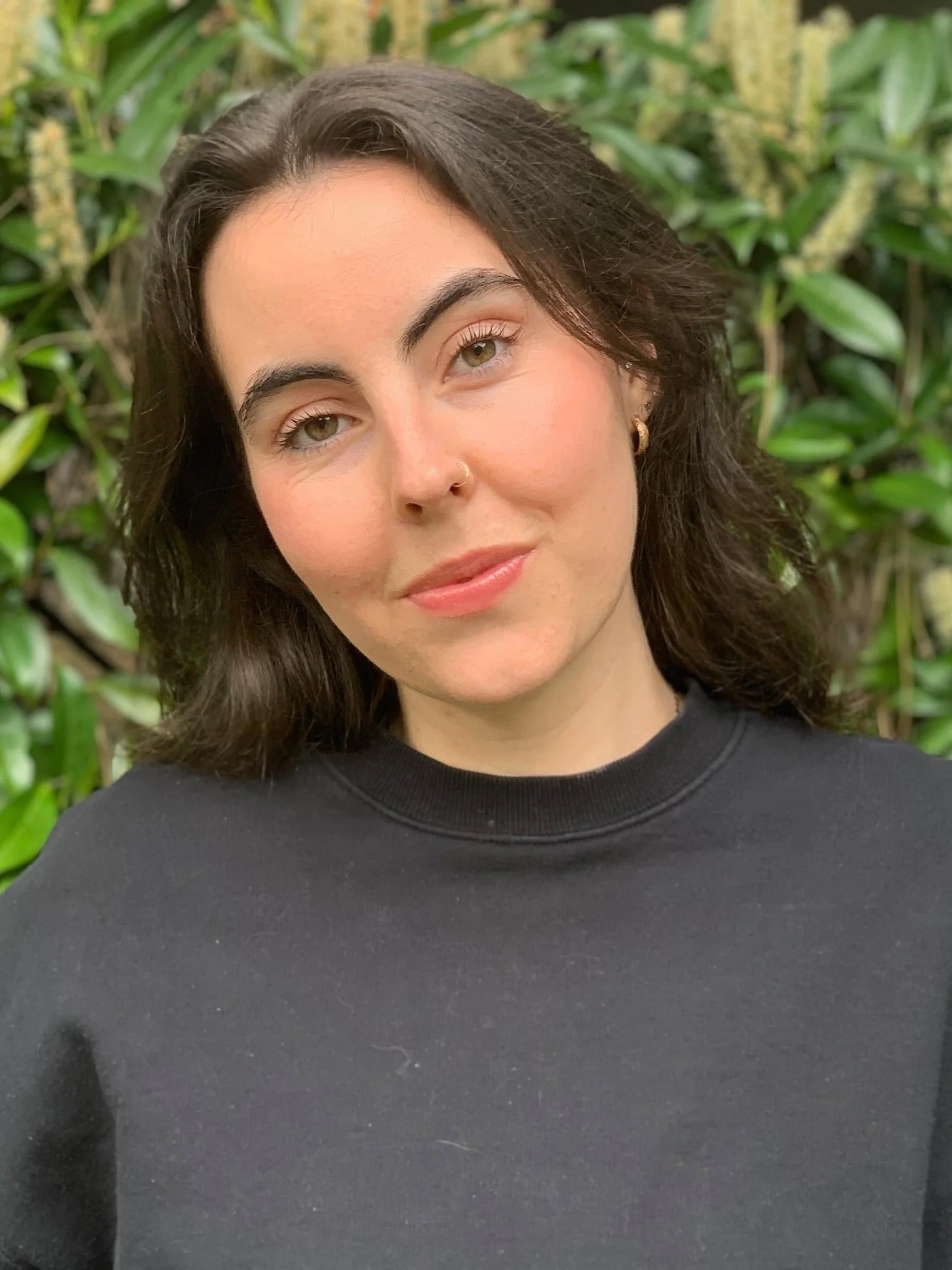 Close-up of a woman with dark brown hair, light makeup, wearing a black shirt, standing in front of green foliage.