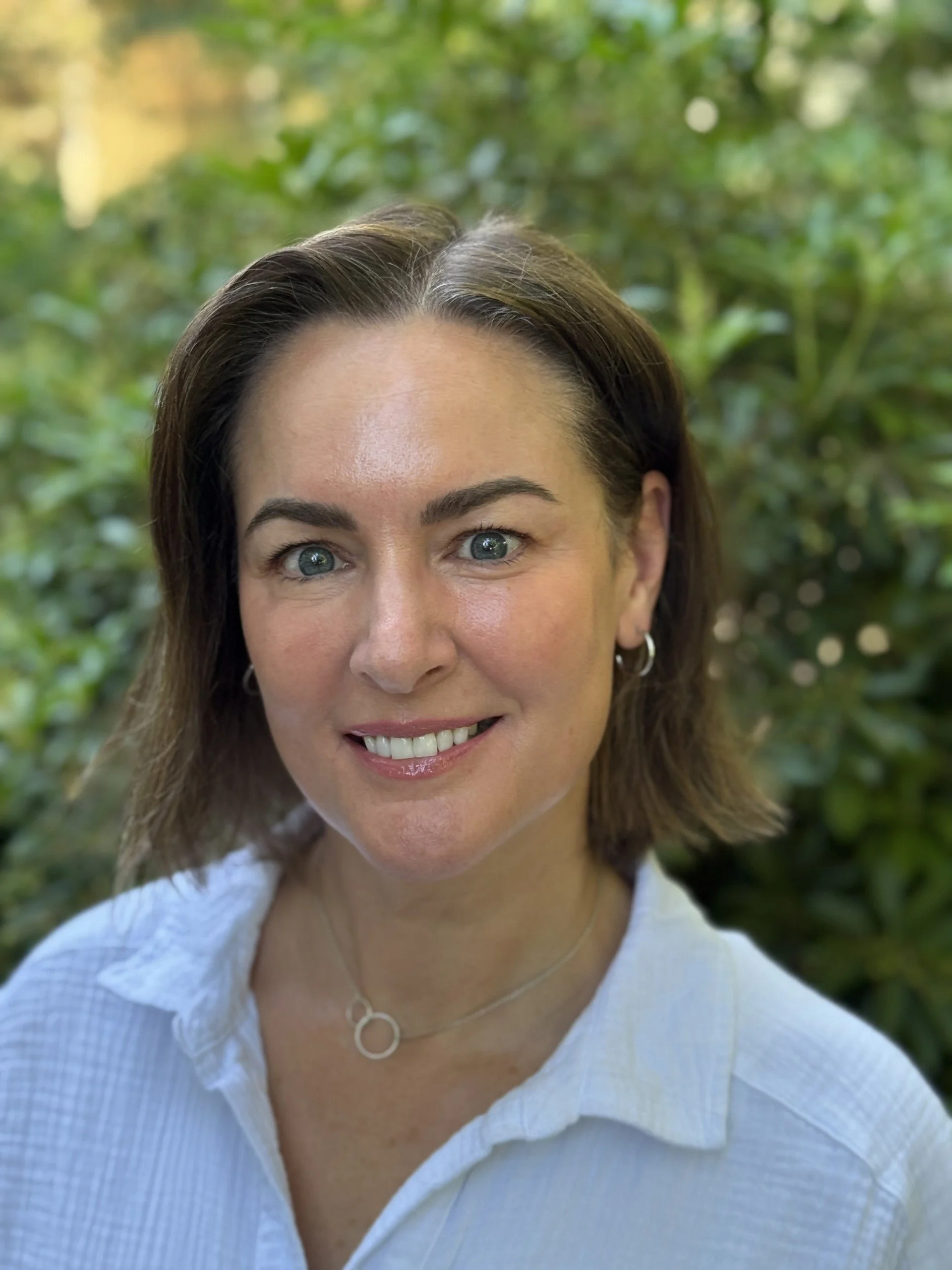 A smiling woman with shoulder-length brown hair, wearing a black blazer over a white top, standing outdoors in front of green leafy bushes.